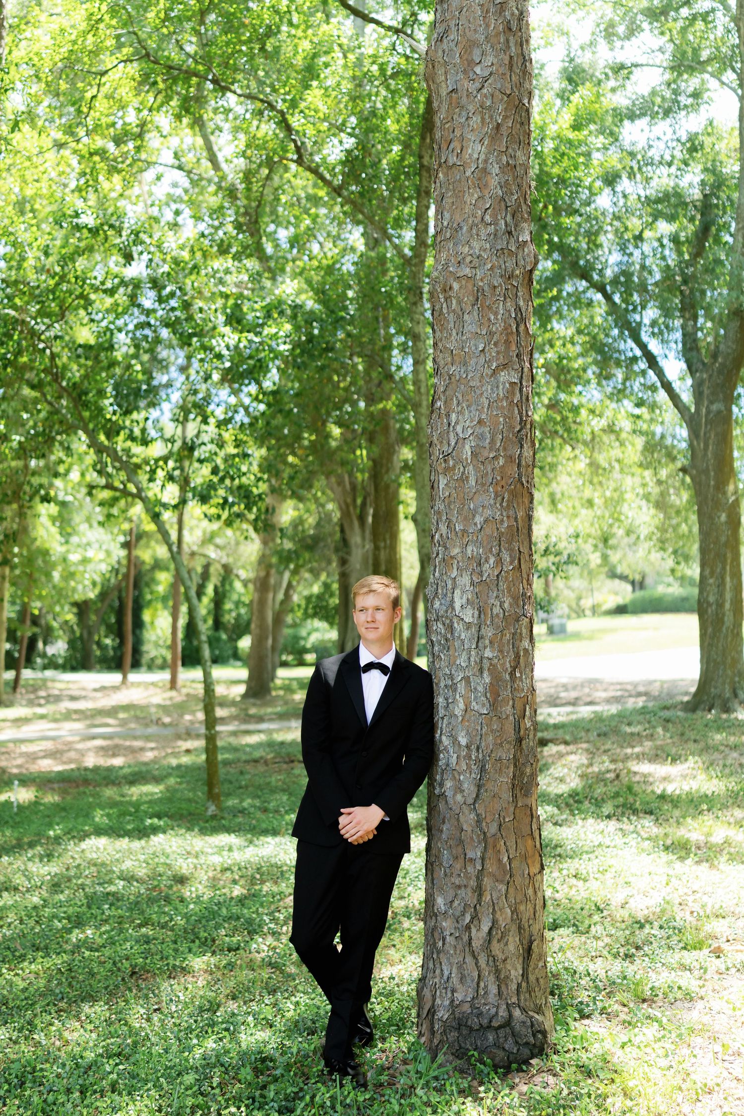 Handsome groom in black tuxedo leaning against a tree trunk in a sunlit green park setting.