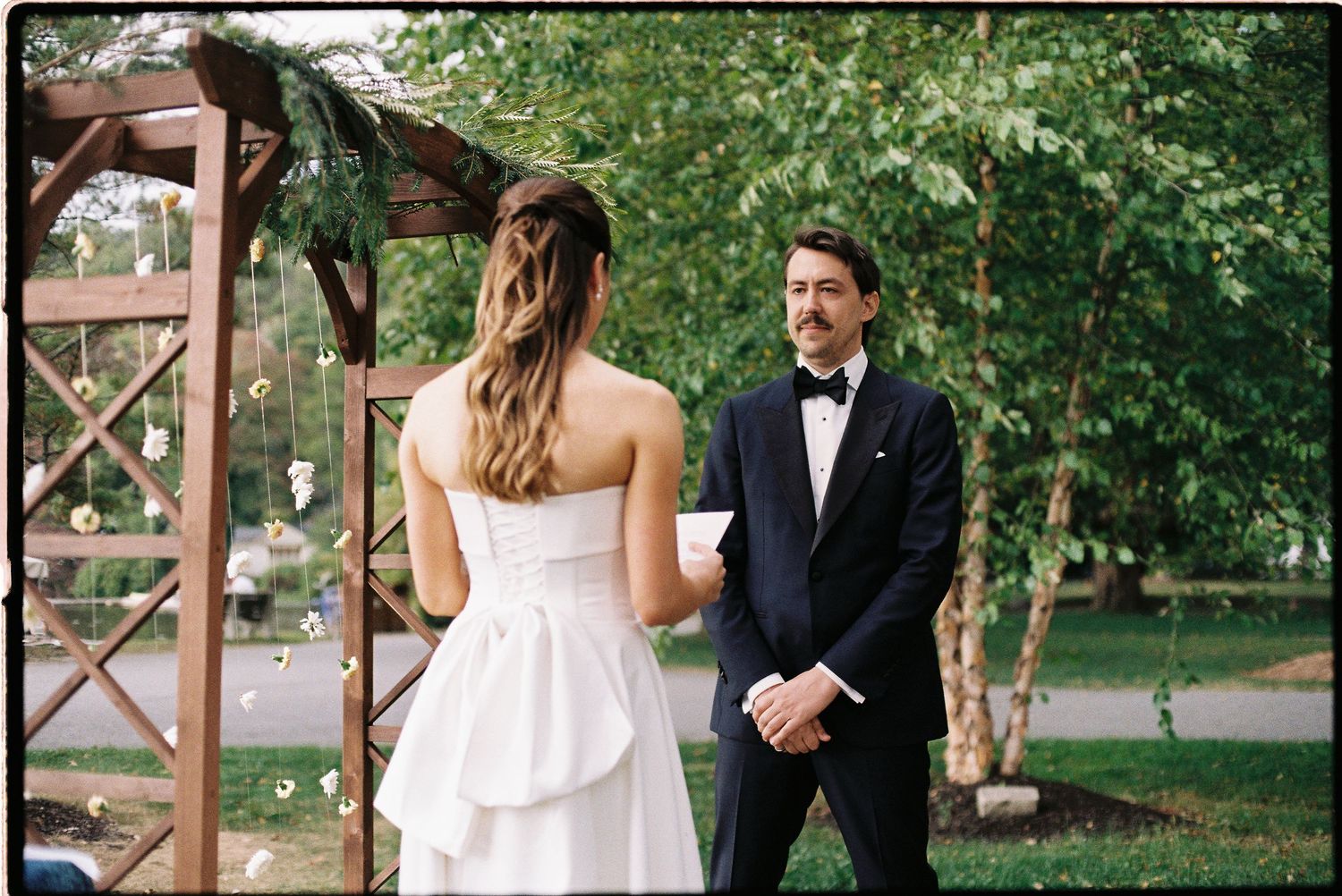 Bride in strapless white gown exchanges vows with groom in black tuxedo at outdoor garden wedding.