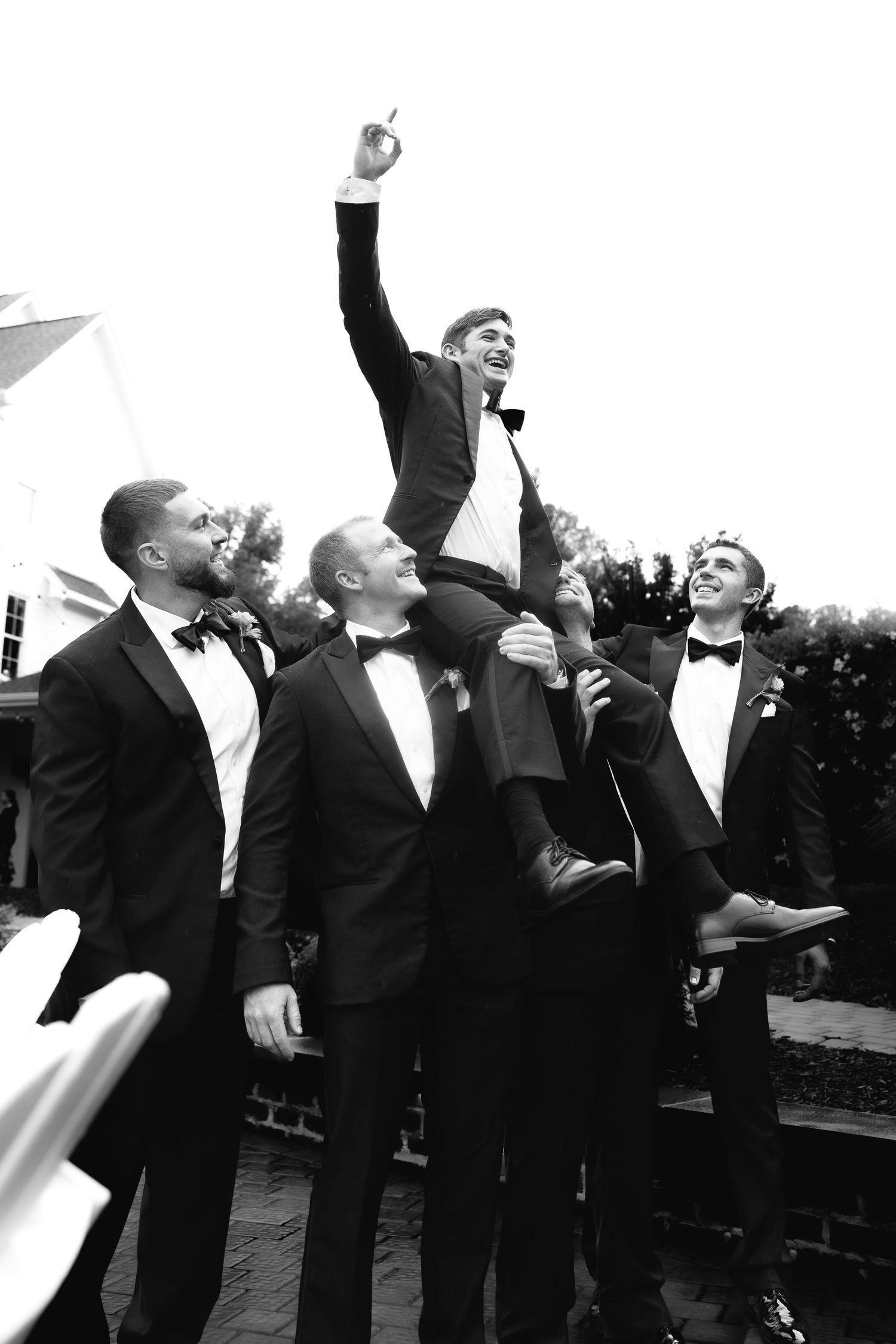 A group of groomsmen in black tuxedos celebrate joyfully outdoors during a wedding celebration.