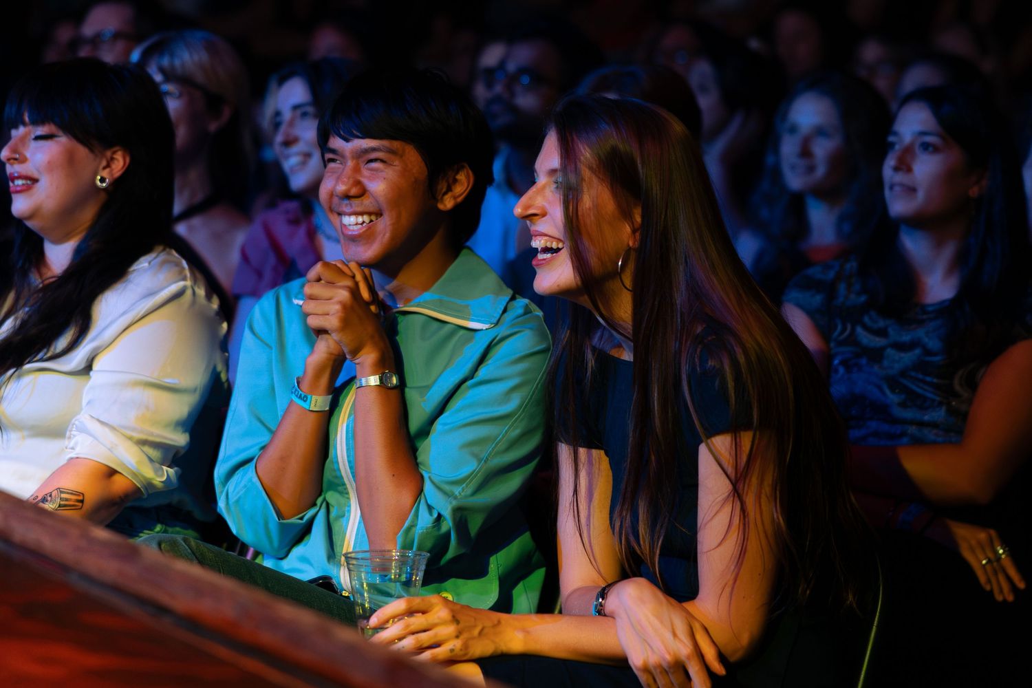 Two laughing friends enjoy a live show together in a crowded, warmly lit theater venue.