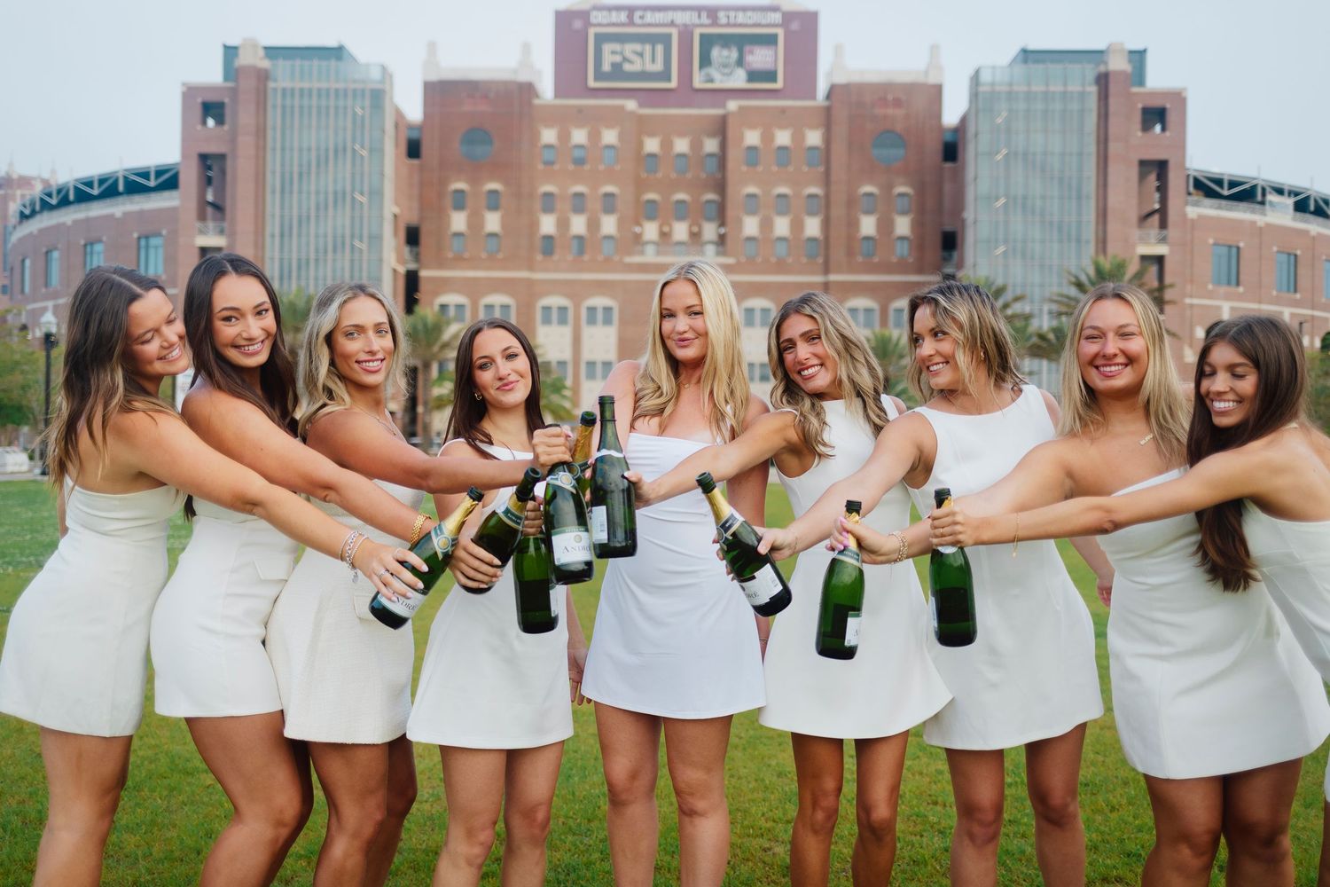 A group in white dresses holds champagne bottles while posing together on grass near brick buildings.