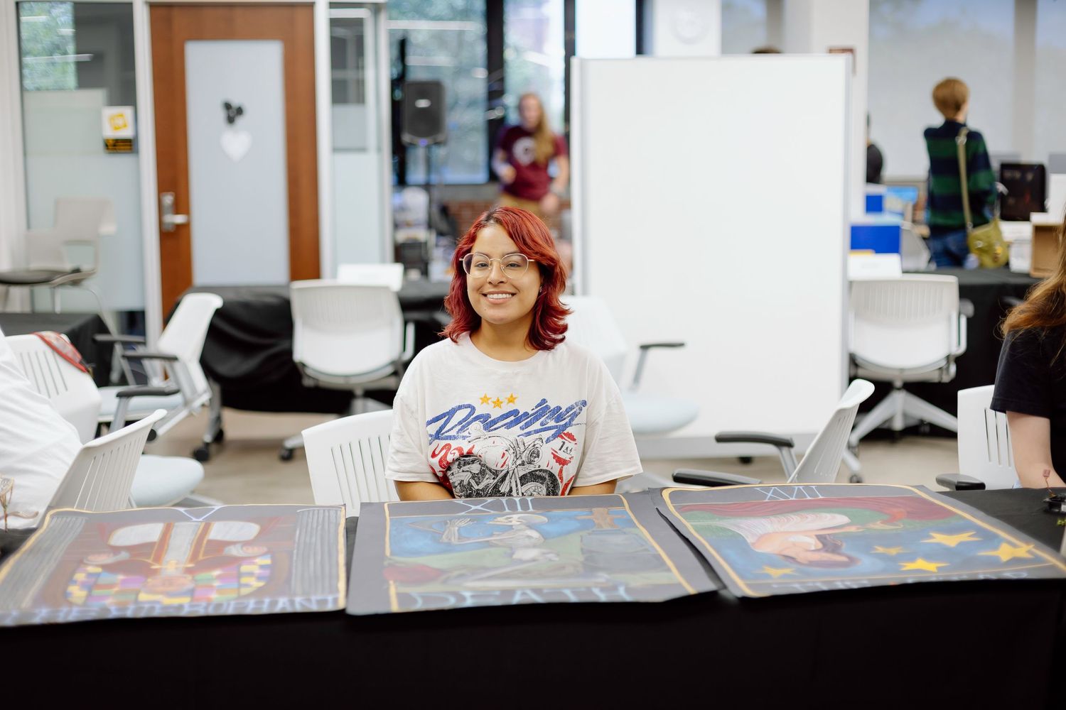 Someone sits smiling at a table displaying photo portfolios at an indoor event or exhibition.