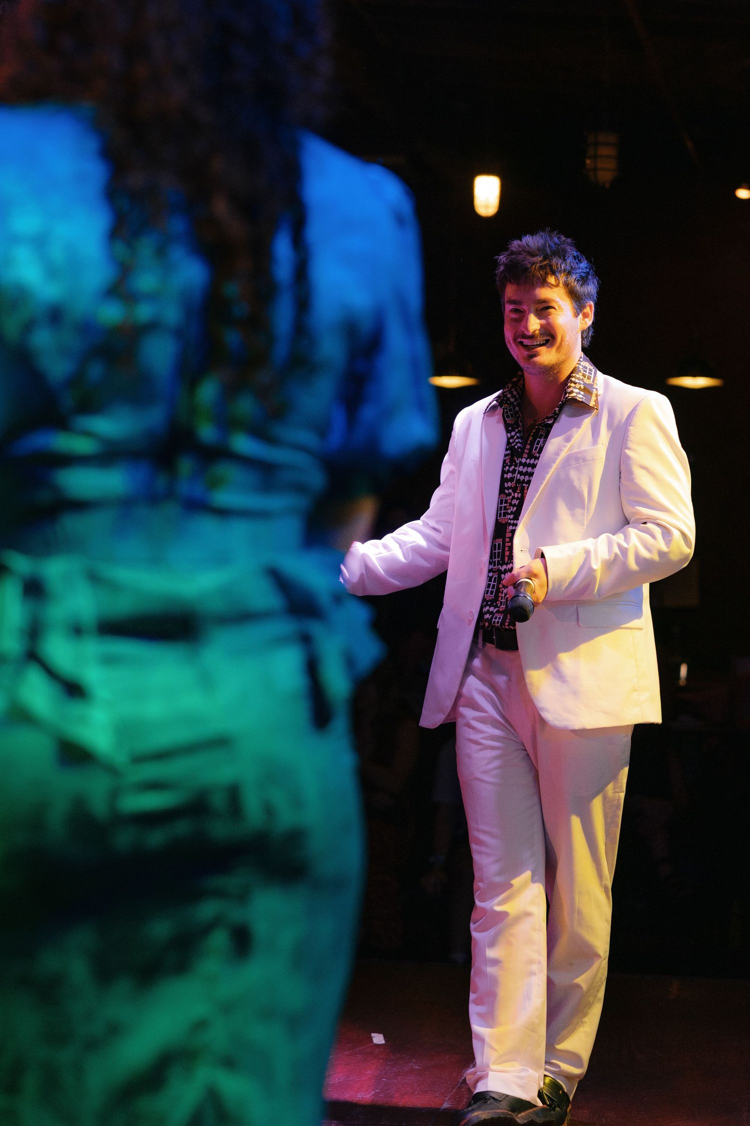 Man in white suit smiling and dancing with a woman in a green dress under colorful stage lighting.