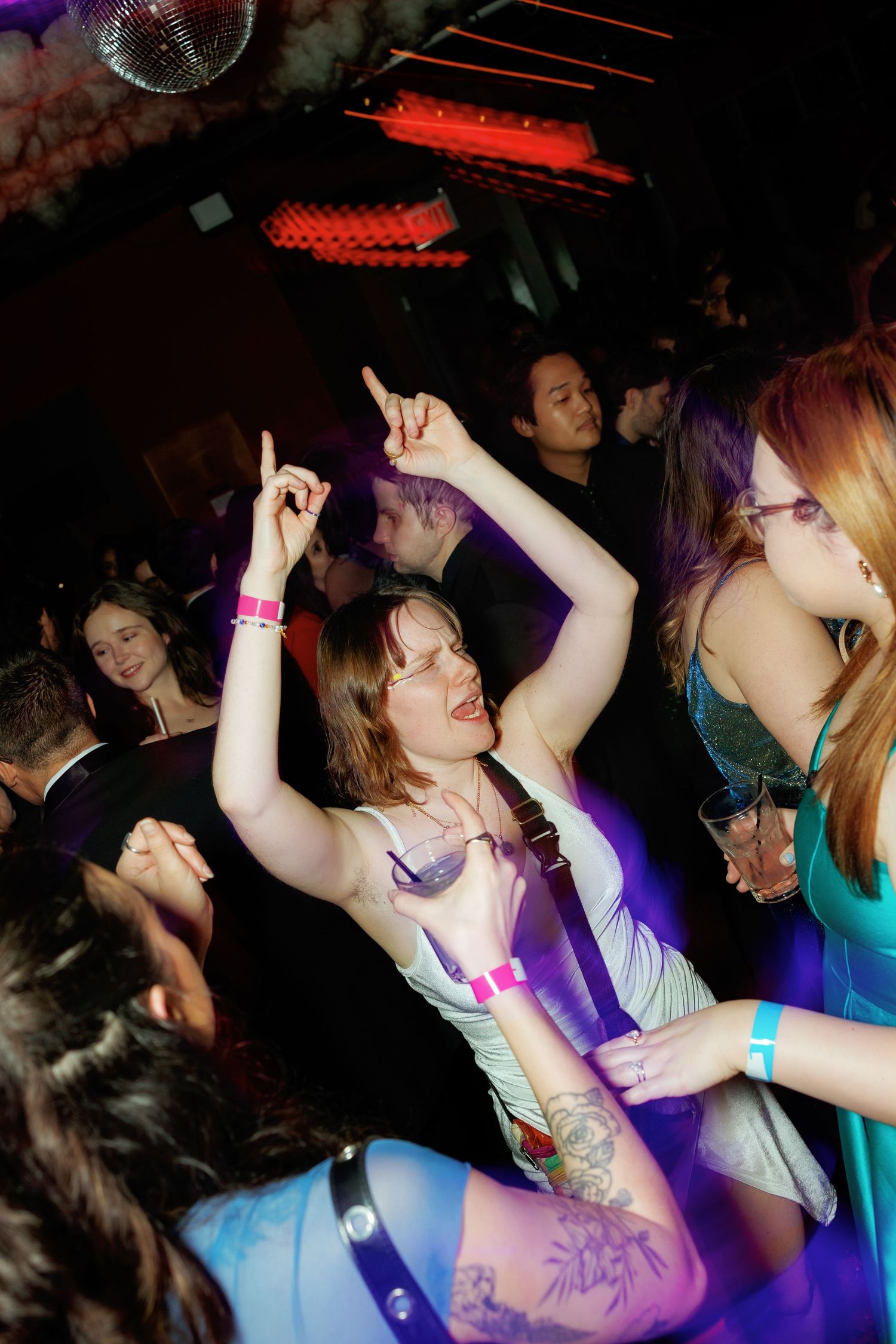 People dancing energetically at a nightclub with disco ball, purple lighting, and colorful wristbands on the dance floor.