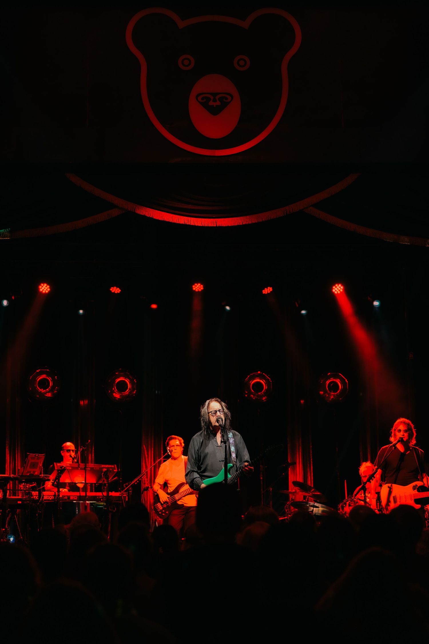 A band performs on stage under dramatic red lighting with a large cartoon bear logo illuminated above them.
