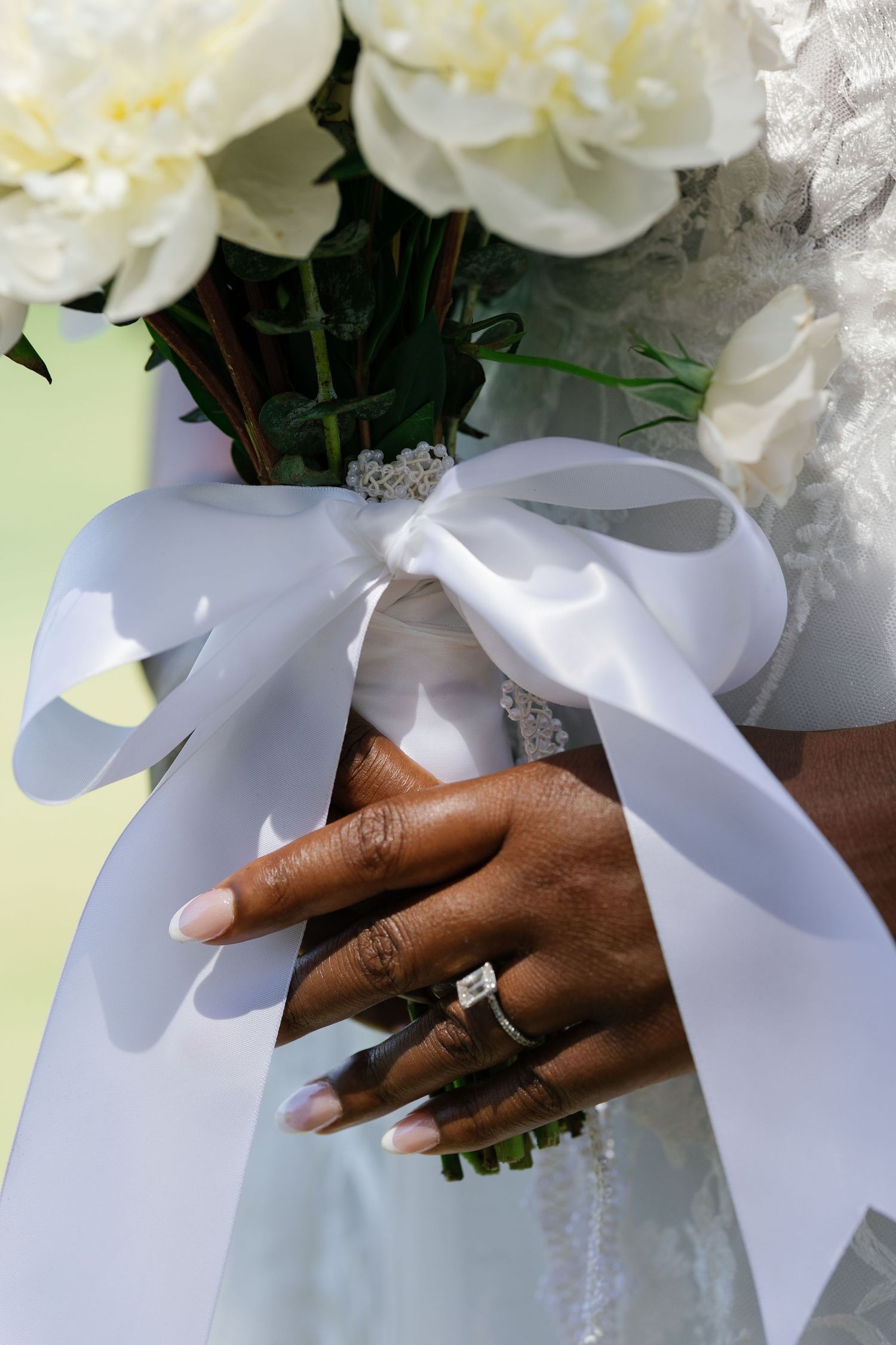 Close-up of bride's hand holding white peony bouquet with satin ribbon, featuring elegant diamond engagement ring.