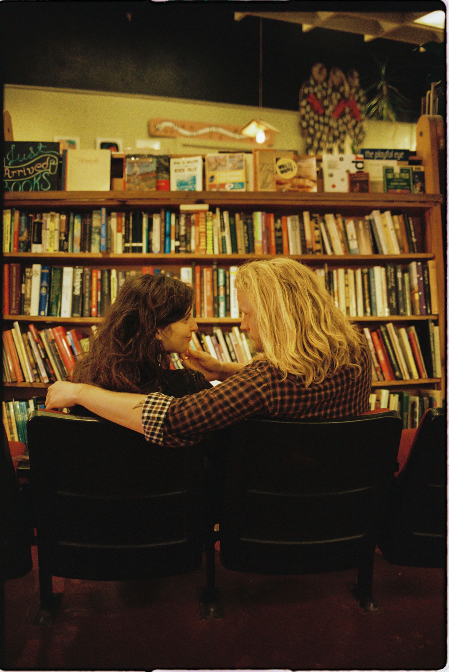 Two women with long hair sit close together in theater-style chairs in front of a tall wooden bookshelf filled with colorful books.