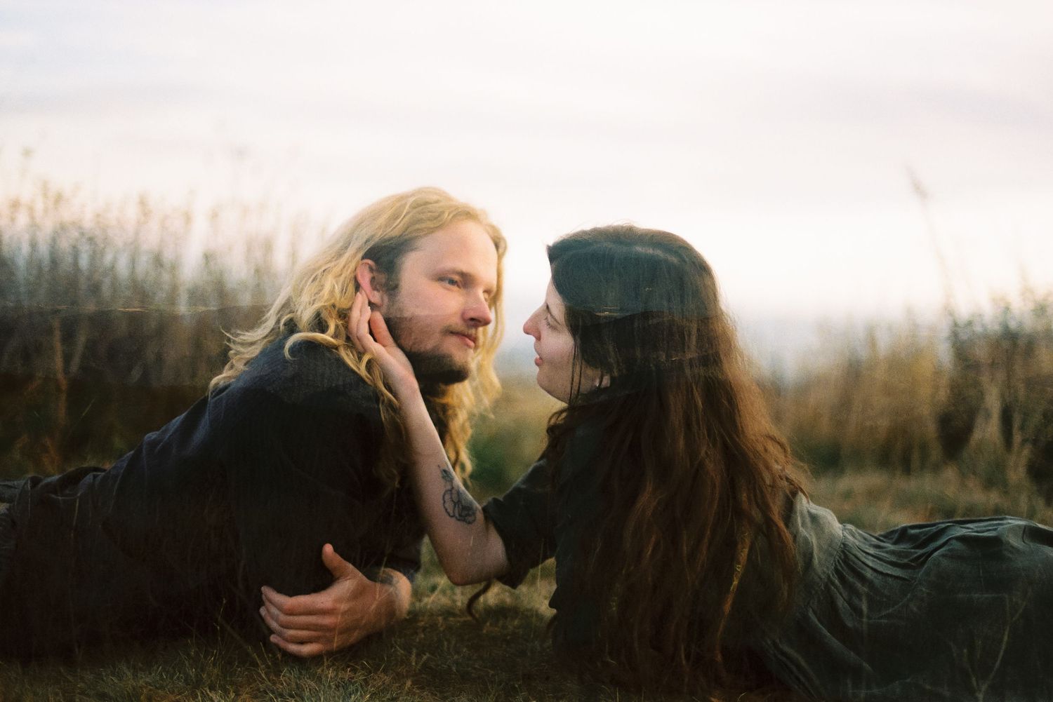 A long-haired man and a dark-haired woman lie facing each other on a grassy hillside at sunset as she gently touches his face.