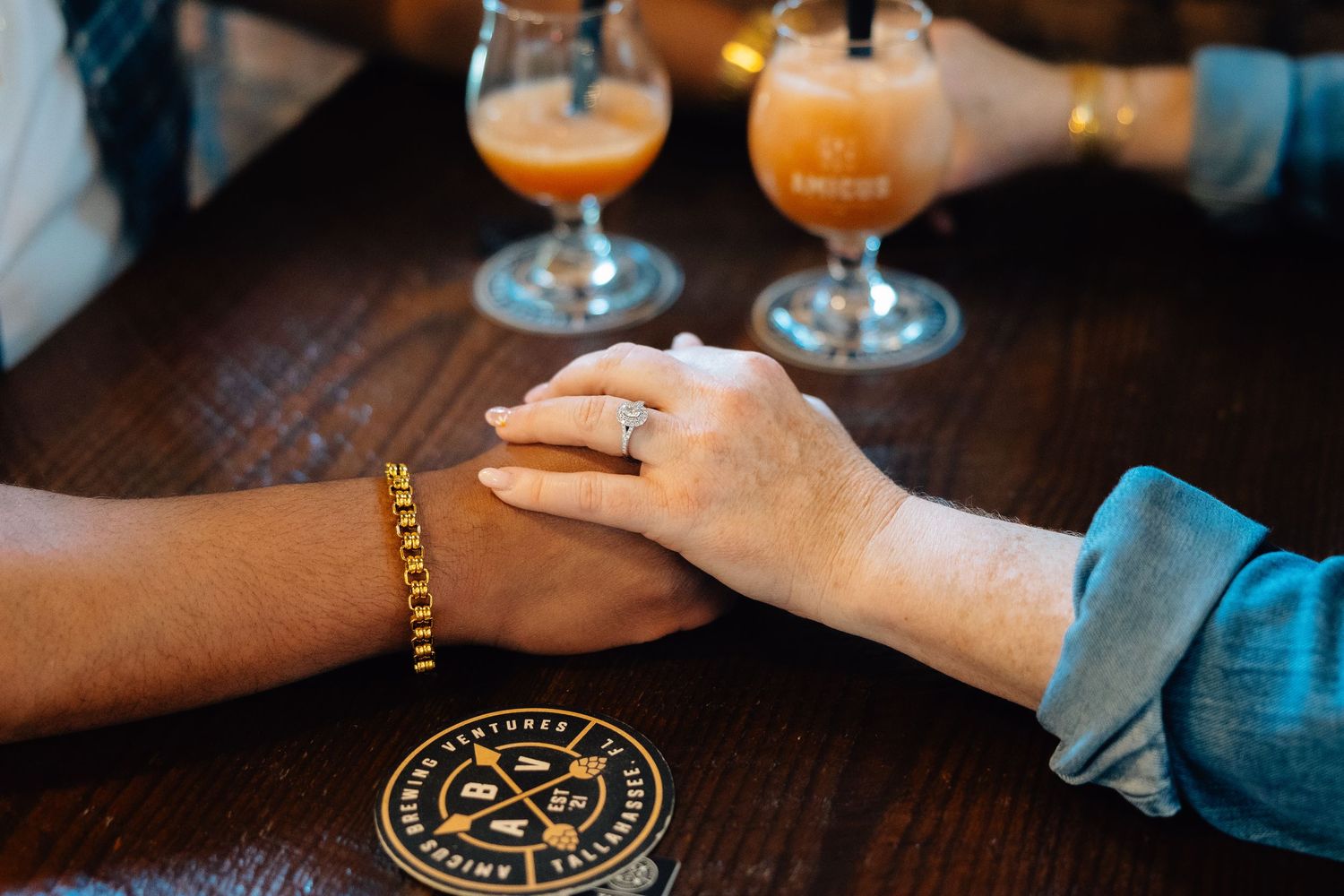 A cocktail glass filled with an orange-colored drink sits on a dark wooden bar, alongside other beverages and hands.