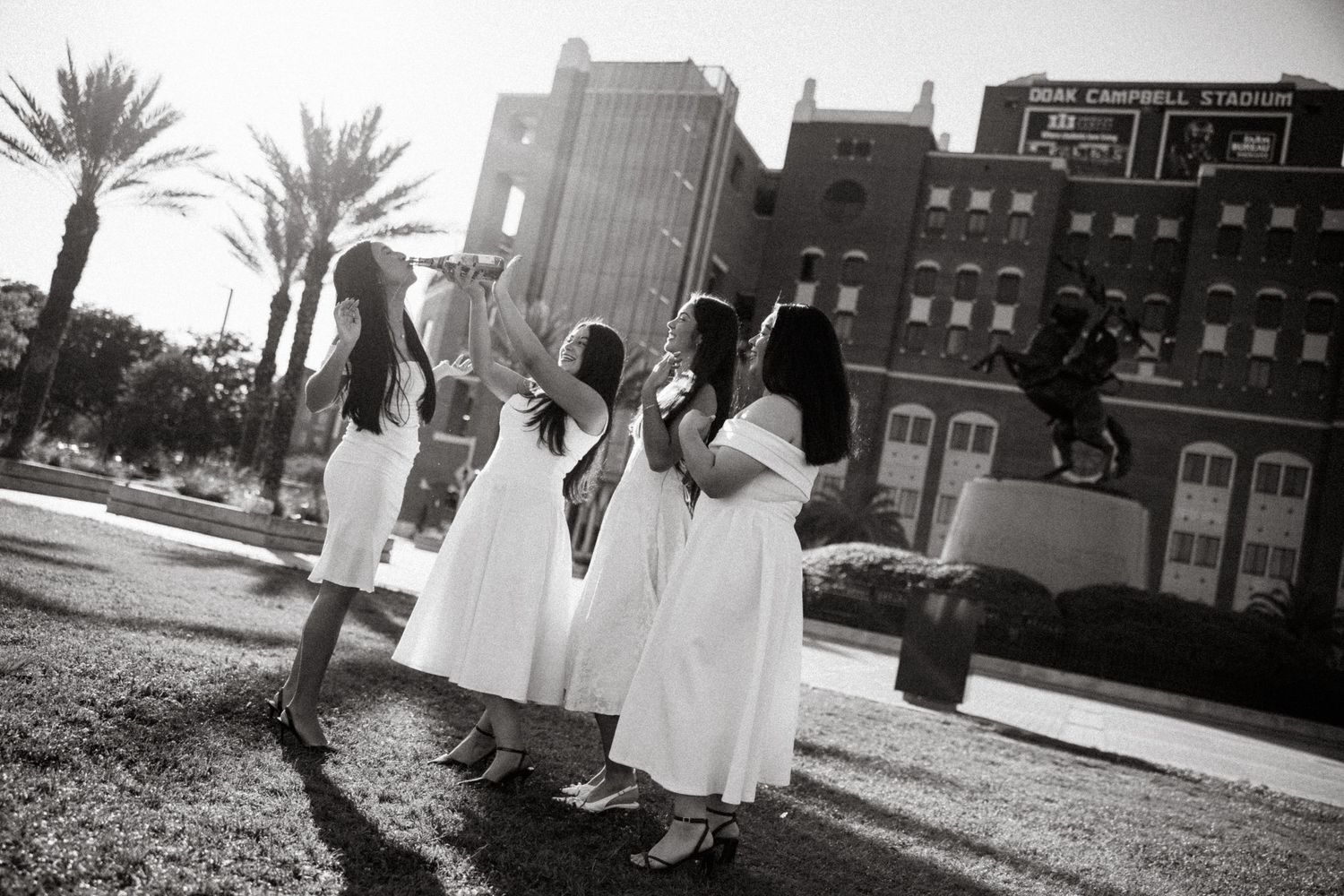Four women in white dresses celebrate outdoors near a stadium, captured in a classic black and white photo.