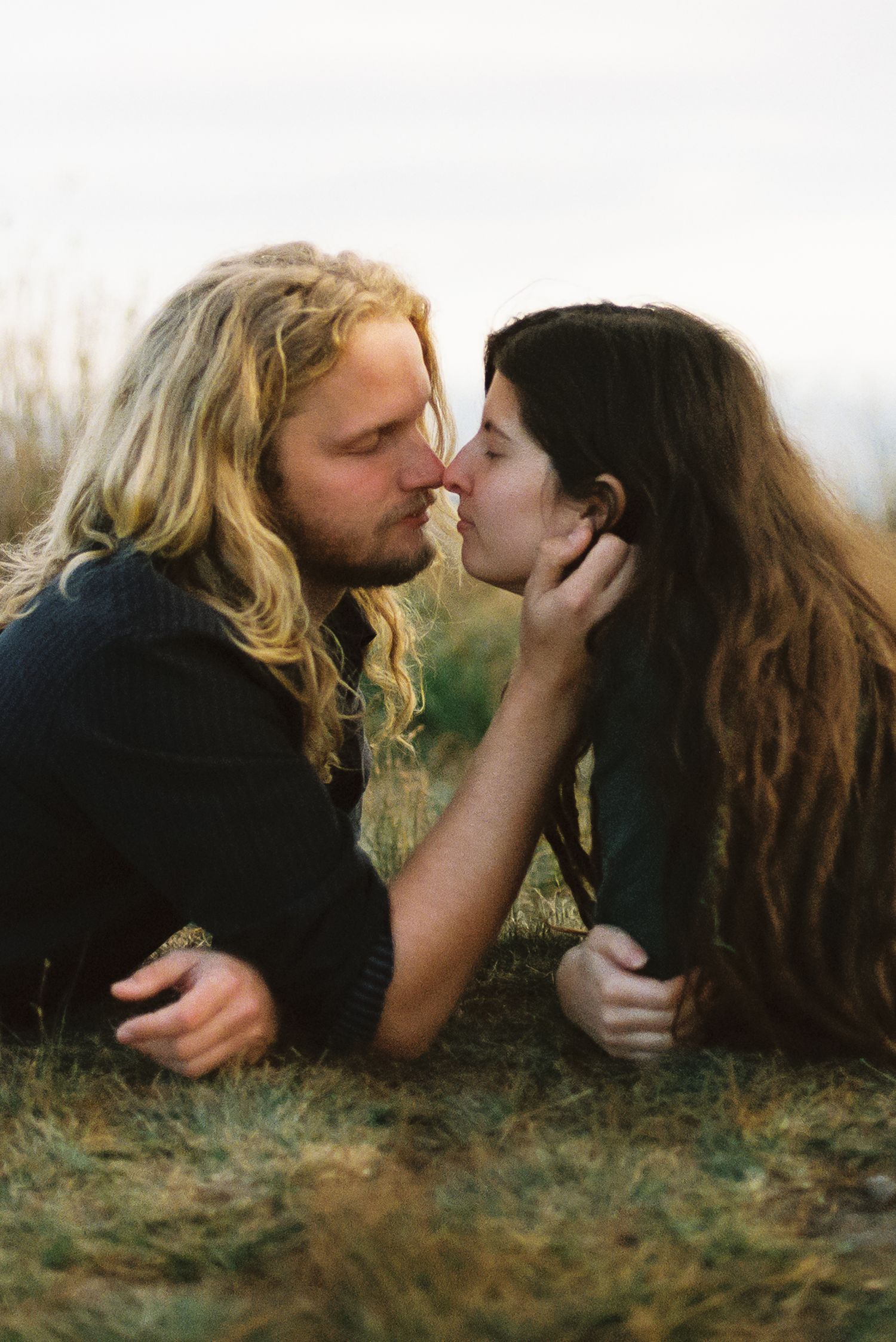 A long-haired couple lies on grass in a romantic moment, noses touching as the woman gently cups the man's face.