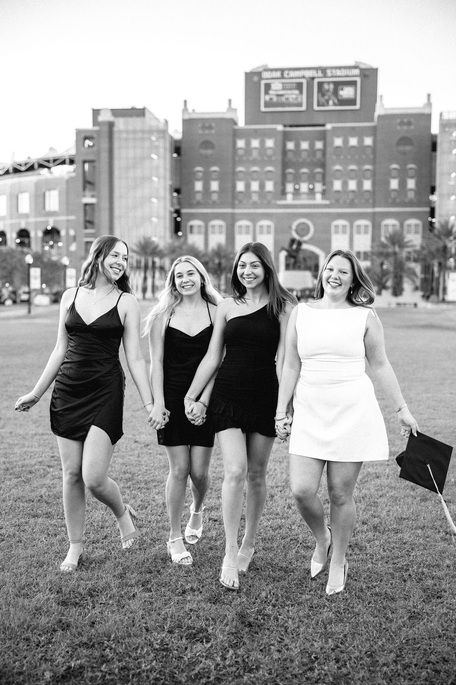 A group of friends in formal dresses walk together on grass with a stadium in the background.