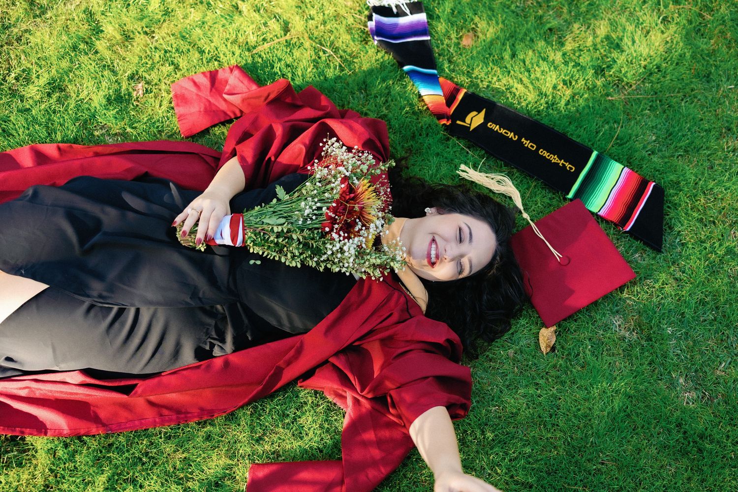 A graduate in red cap and gown lies on green grass in a celebratory pose during graduation.