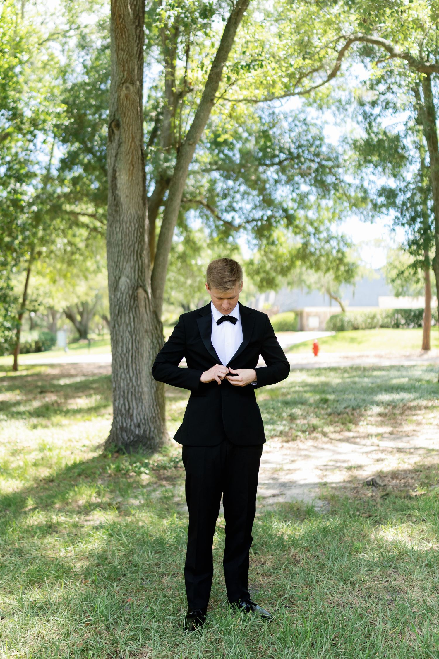 Dapper groom in black tuxedo and bow tie buttoning his jacket while standing outdoors among green trees.
