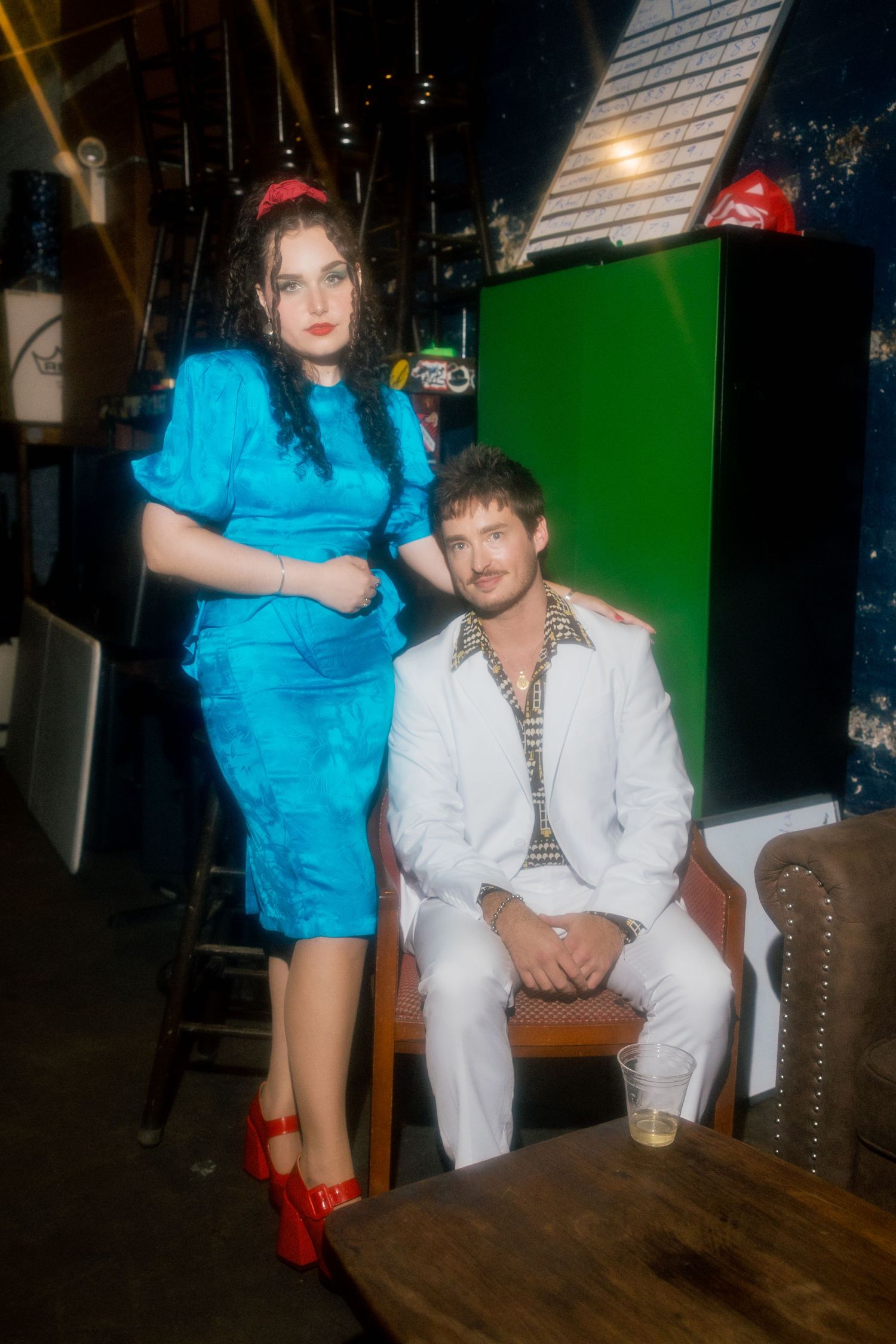 A woman in blue dress and red heels stands beside a man in white suit seated in a retro-styled venue.