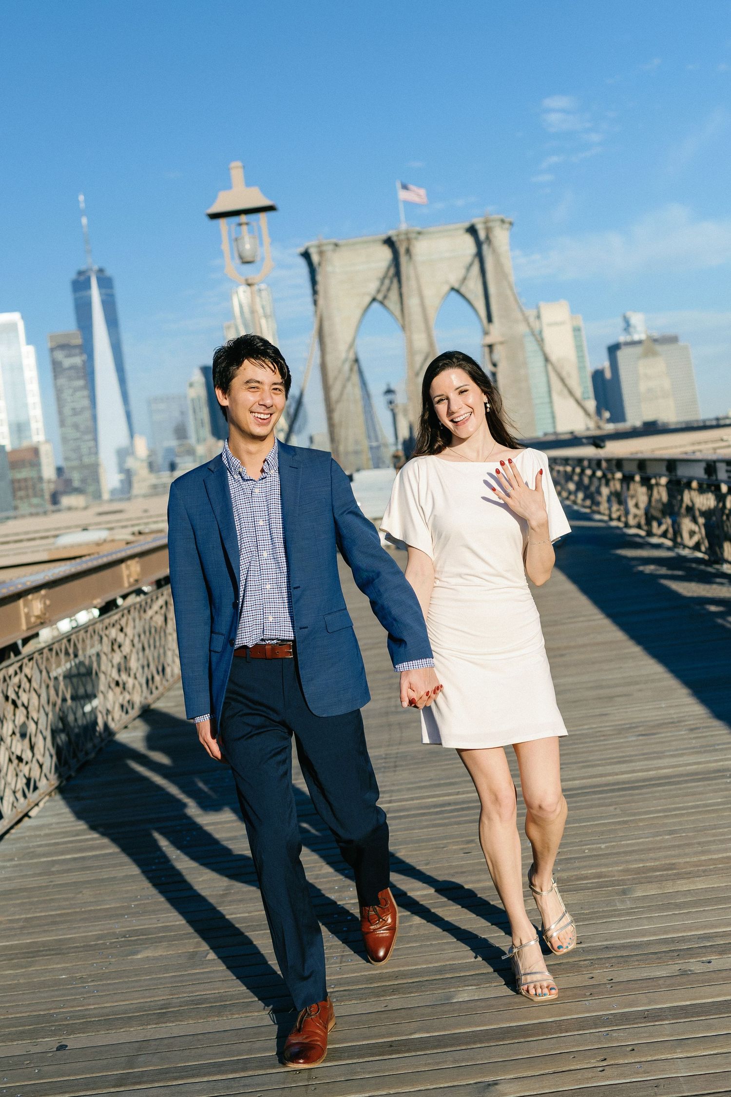 Smiling couple holds hands while walking on the Brooklyn Bridge wooden promenade, with the New York City skyline behind them.