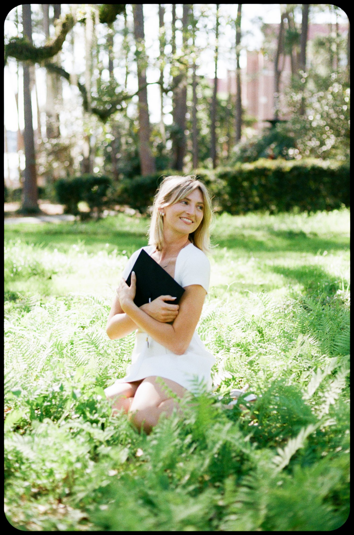 Smiling blonde woman kneeling in lush green garden holding black book, wearing white dress among ferns and trees.