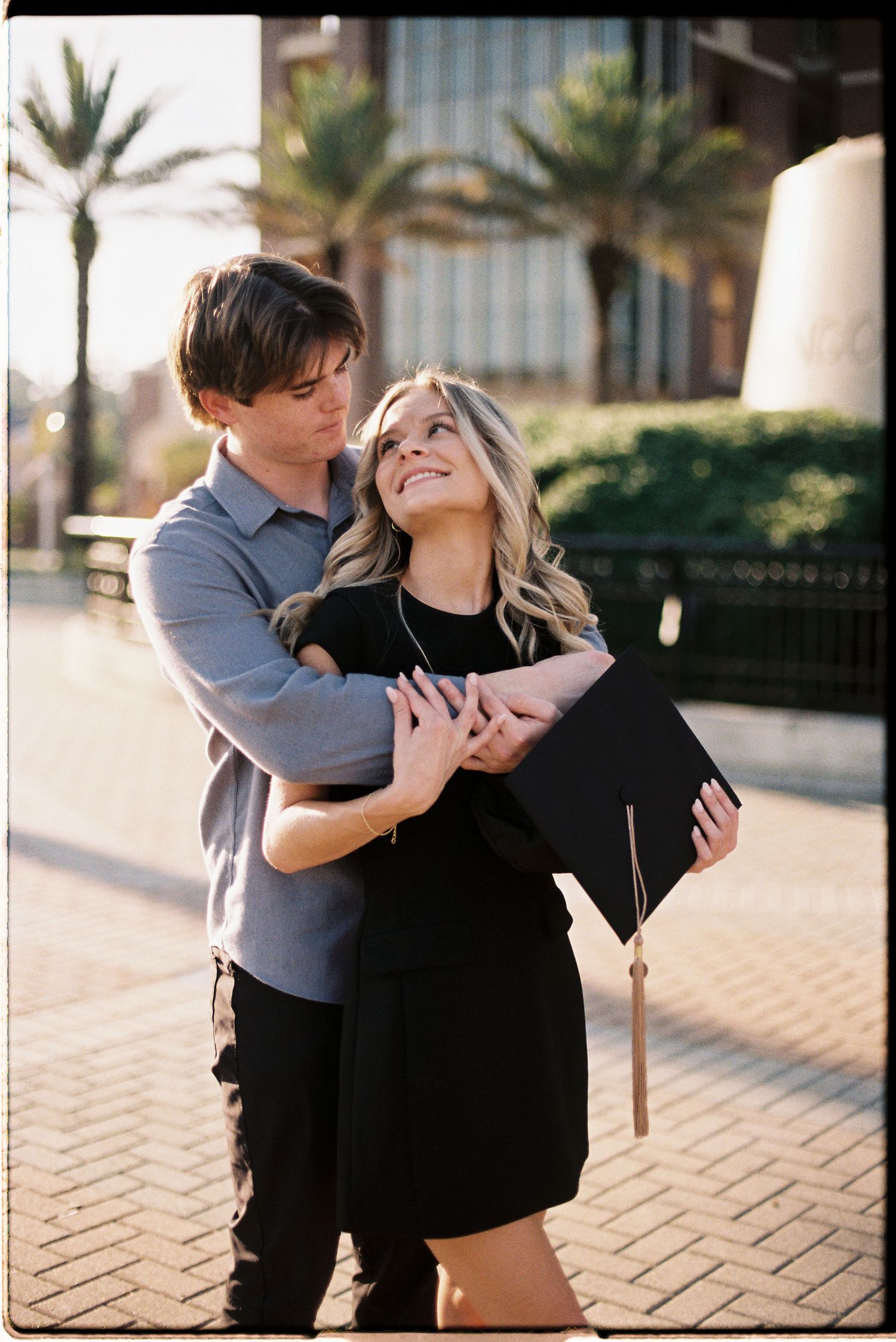 A couple embraces on a brick walkway with palm trees in the background as they celebrate graduation.
