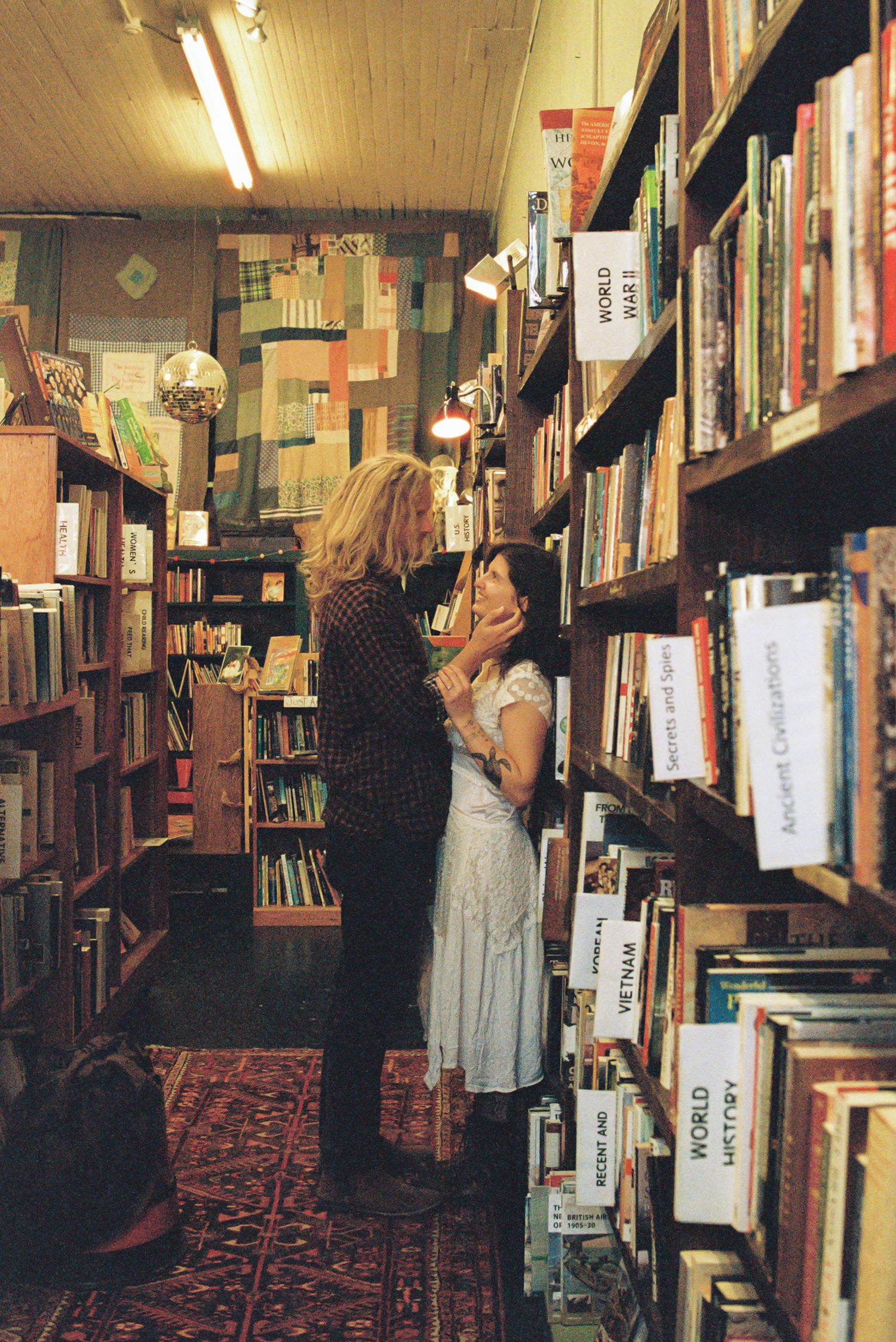Couple embracing in a cozy secondhand bookstore aisle lined with wooden shelves of books, with a red patterned rug and warm lighting.