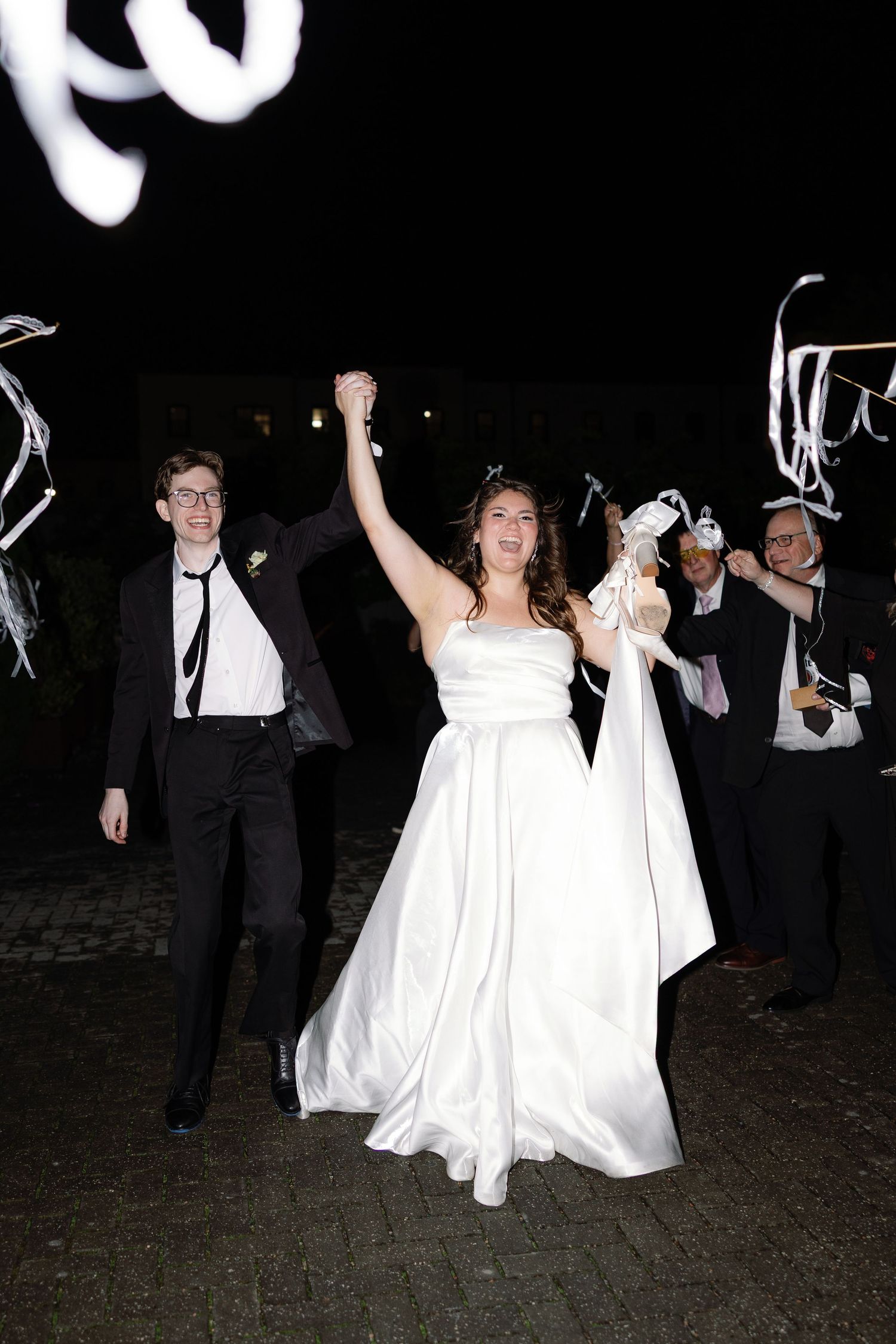 Joyful bride in white gown raises hand with groom in tuxedo during outdoor nighttime wedding celebration.