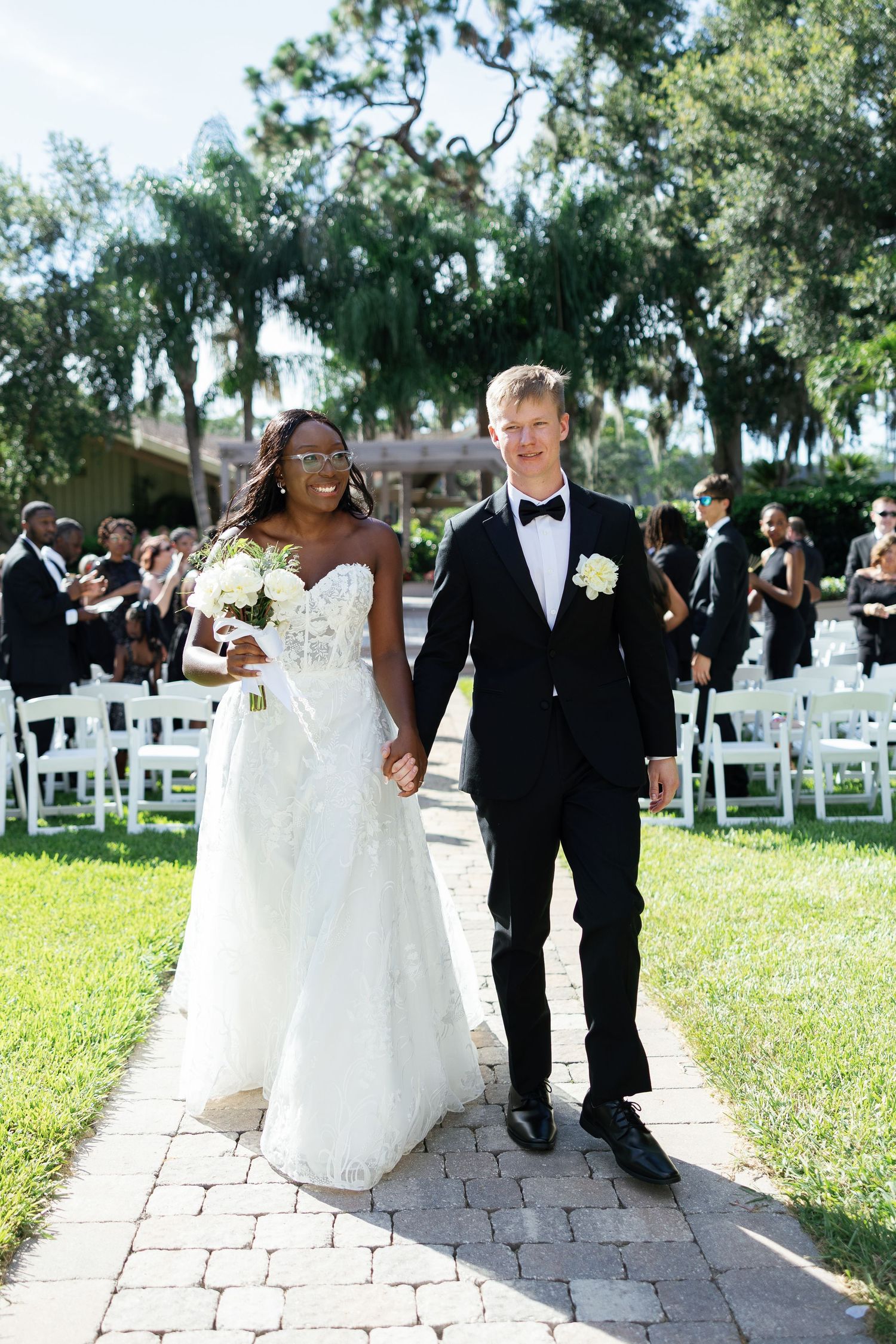 Interracial couple walks down wedding aisle, bride in lace gown holding white bouquet, groom in black tuxedo.
