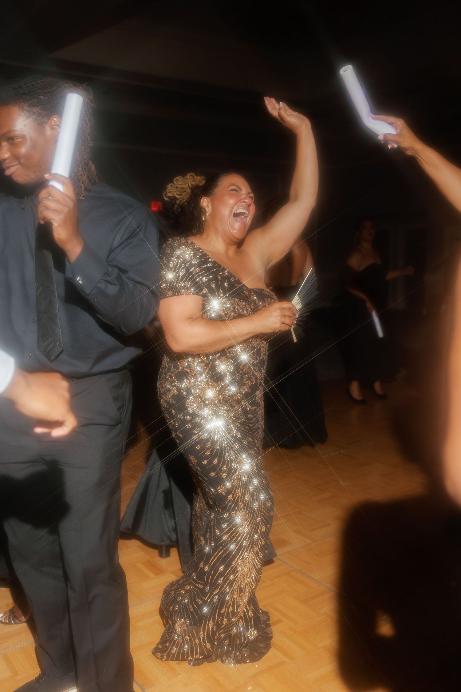 Woman in sparkling black gown dances joyfully with glow stick at lively wedding reception celebration.