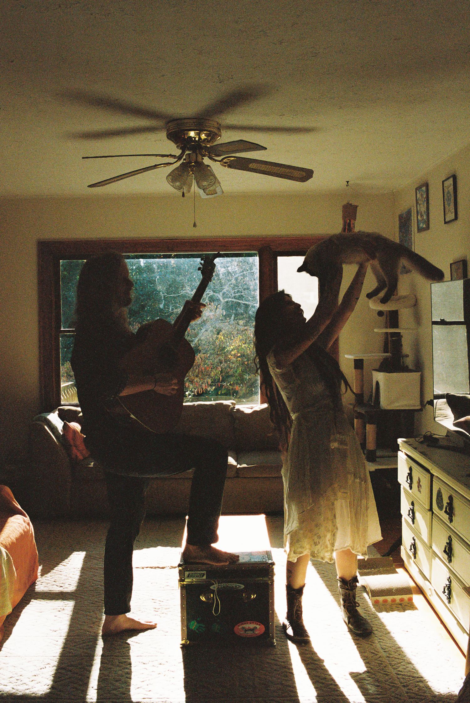 Two people in a sunlit living room under a ceiling fan, one playing acoustic guitar while the other lifts a cat overhead.