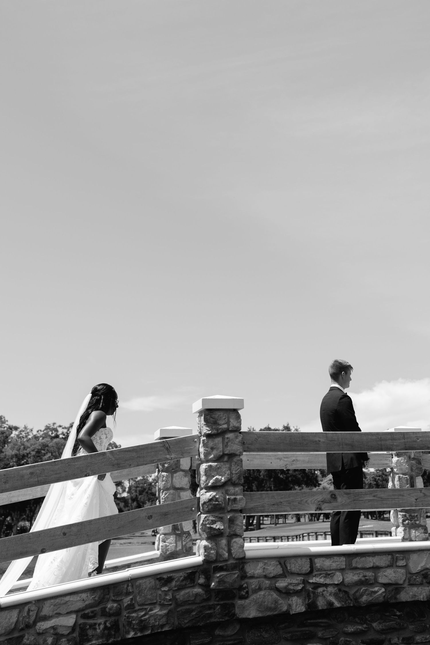 Black and white photo of bride and groom on a stone bridge, standing apart in a romantic first look moment.