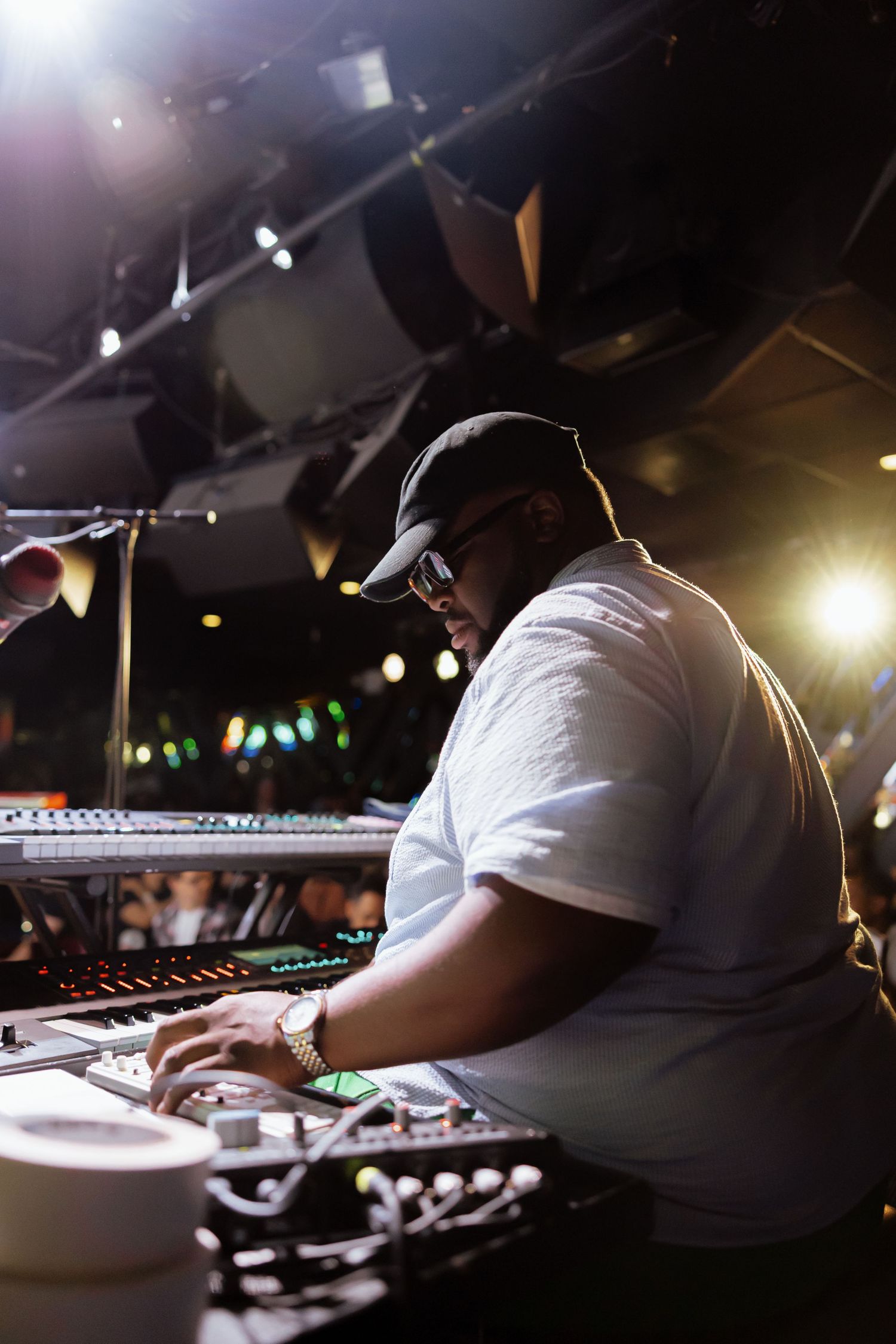 DJ mixing music at soundboard under stage lights in dark concert venue wearing white shirt and cap.
