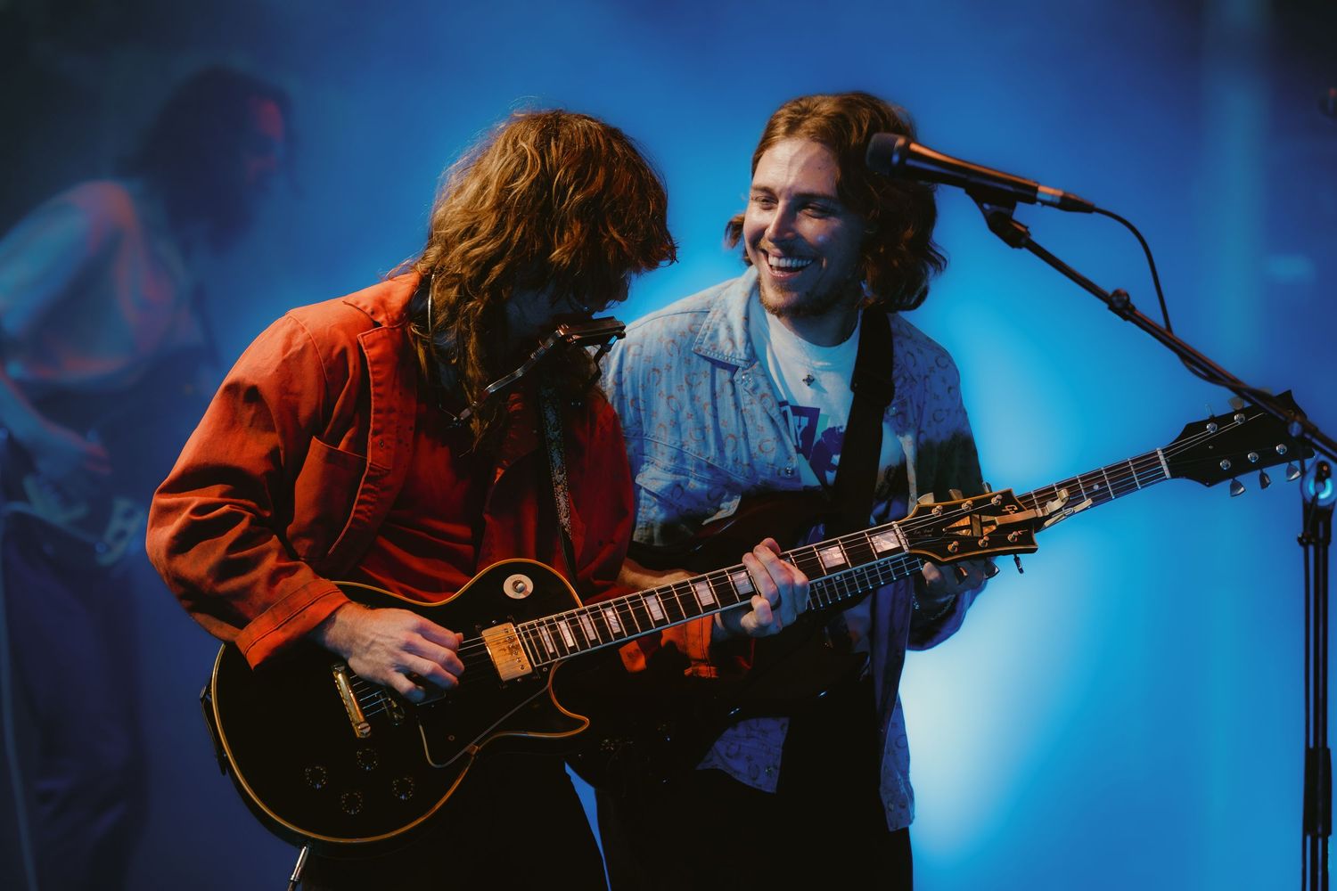 Two guitarists performing on stage with blue lighting, one in red jacket playing a Les Paul guitar.