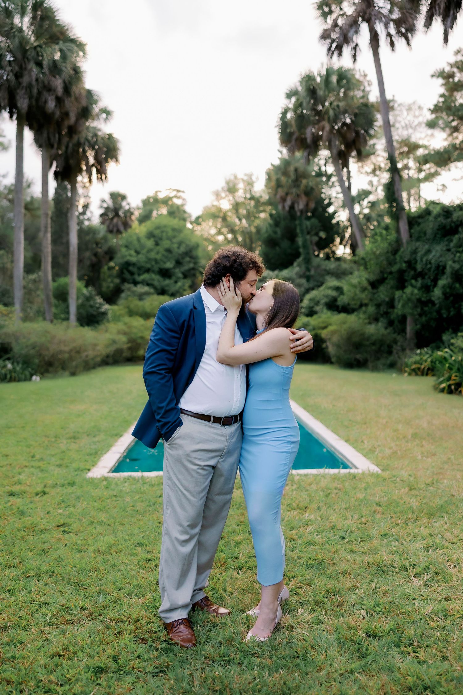 Three people stand together on a grassy lawn with palm trees in the background, sharing a tender moment.