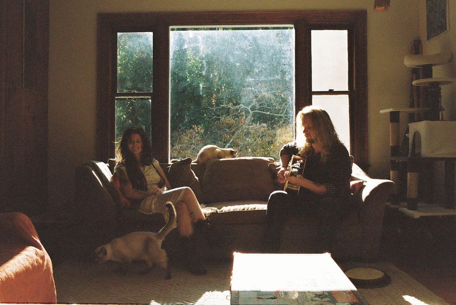 Two women relax on a brown couch in a sunlit living room with a large window, while a small dog sits on the floor nearby.