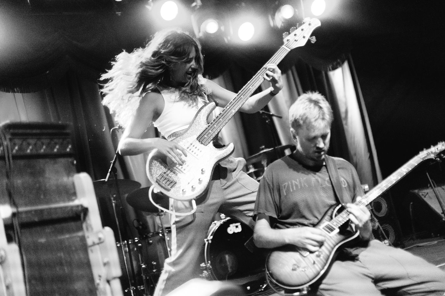 Black and white concert photo showing musicians performing with guitars on stage under bright spotlights.