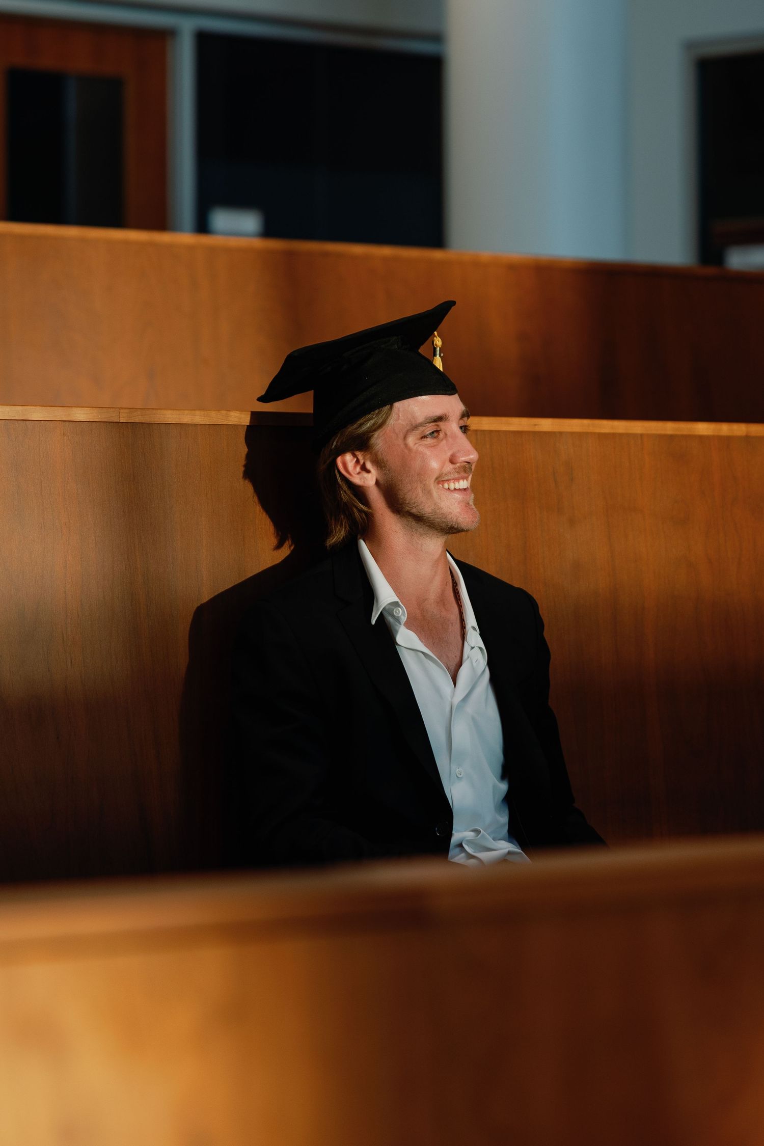 Smiling male graduate wearing black cap and blazer sitting in wooden auditorium seating during graduation ceremony.