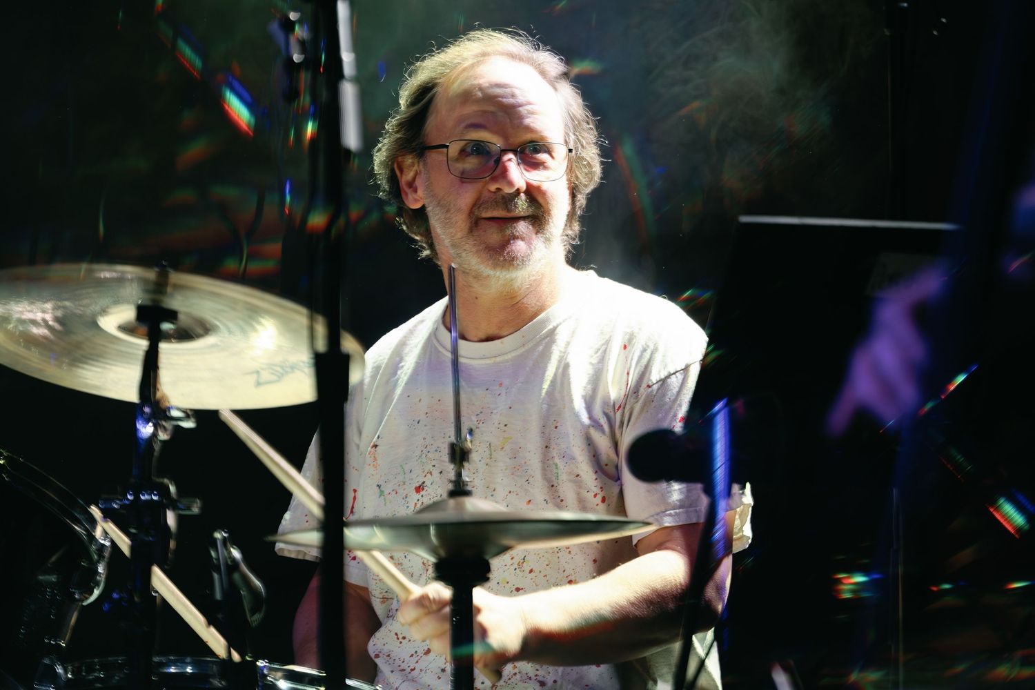 A drummer performs behind a drum kit in a white t-shirt with dramatic lighting and lens flares in the background.