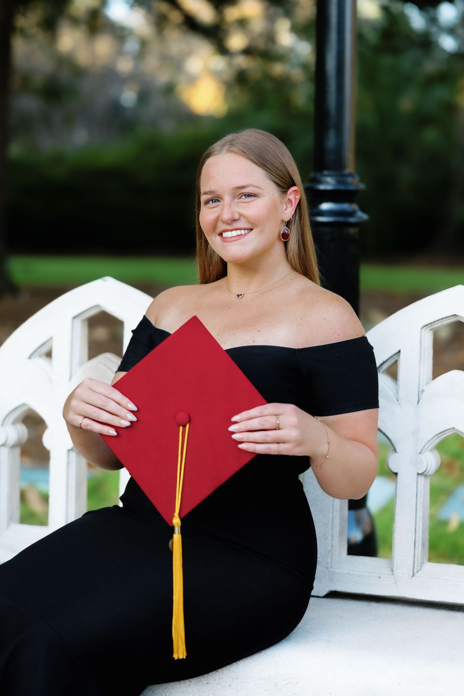 Graduate in black off-shoulder dress sits on white bench holding red diploma and gold tassel during outdoor photo session.