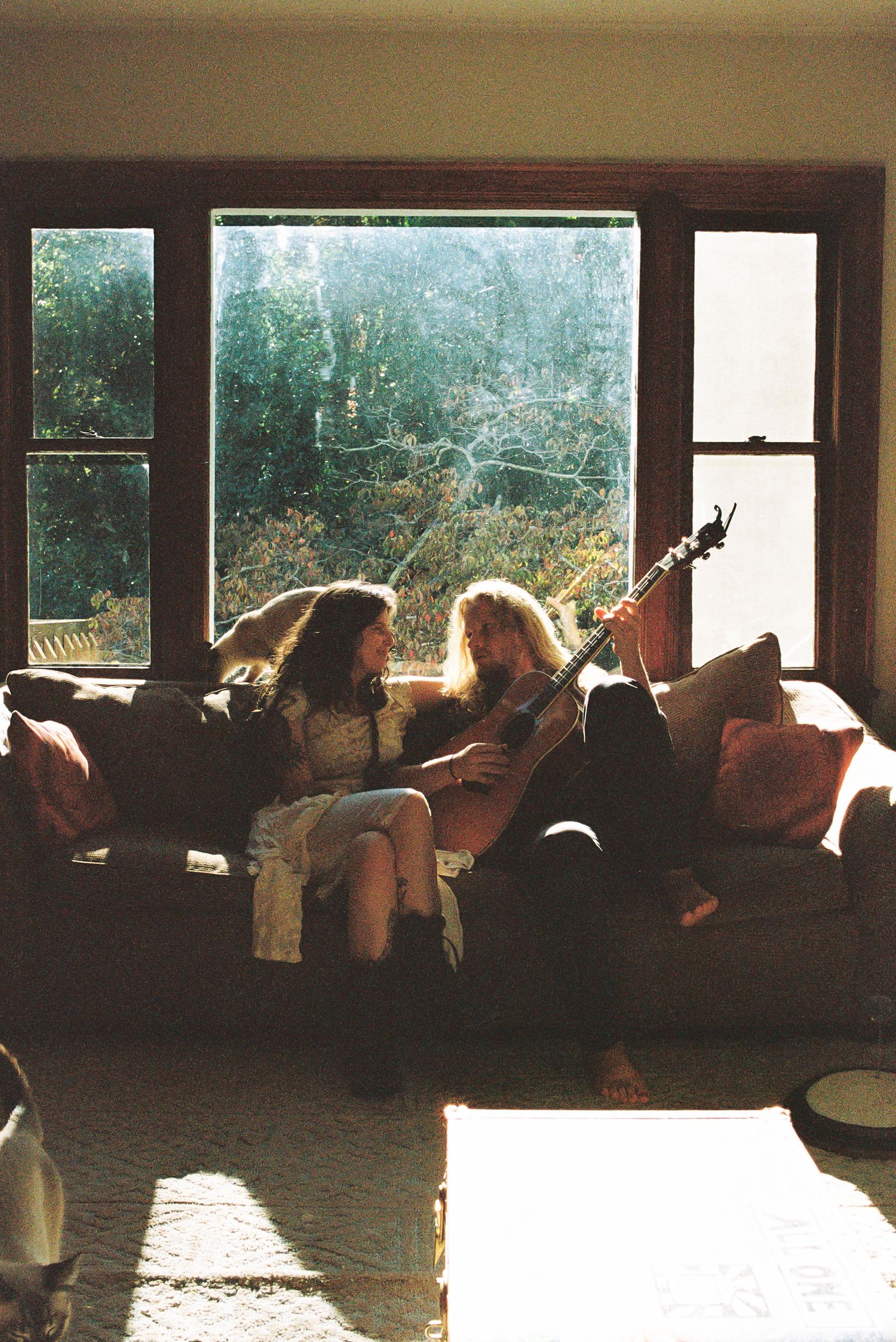 Two women sit close on a couch playing an acoustic guitar together in front of a sunlit window overlooking green trees.