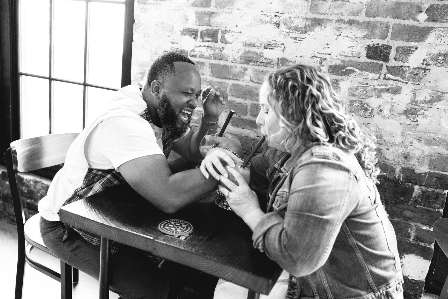 Black and white photo of two people sitting at a table engaged in an arm wrestling match against a brick wall background.