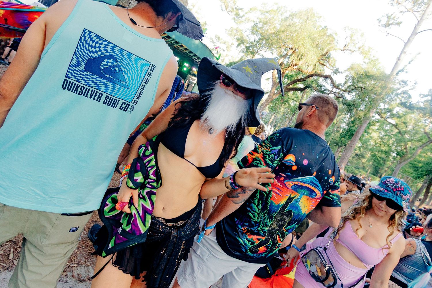 Festival attendees in colorful attire gather outdoors at a summer music event among trees.