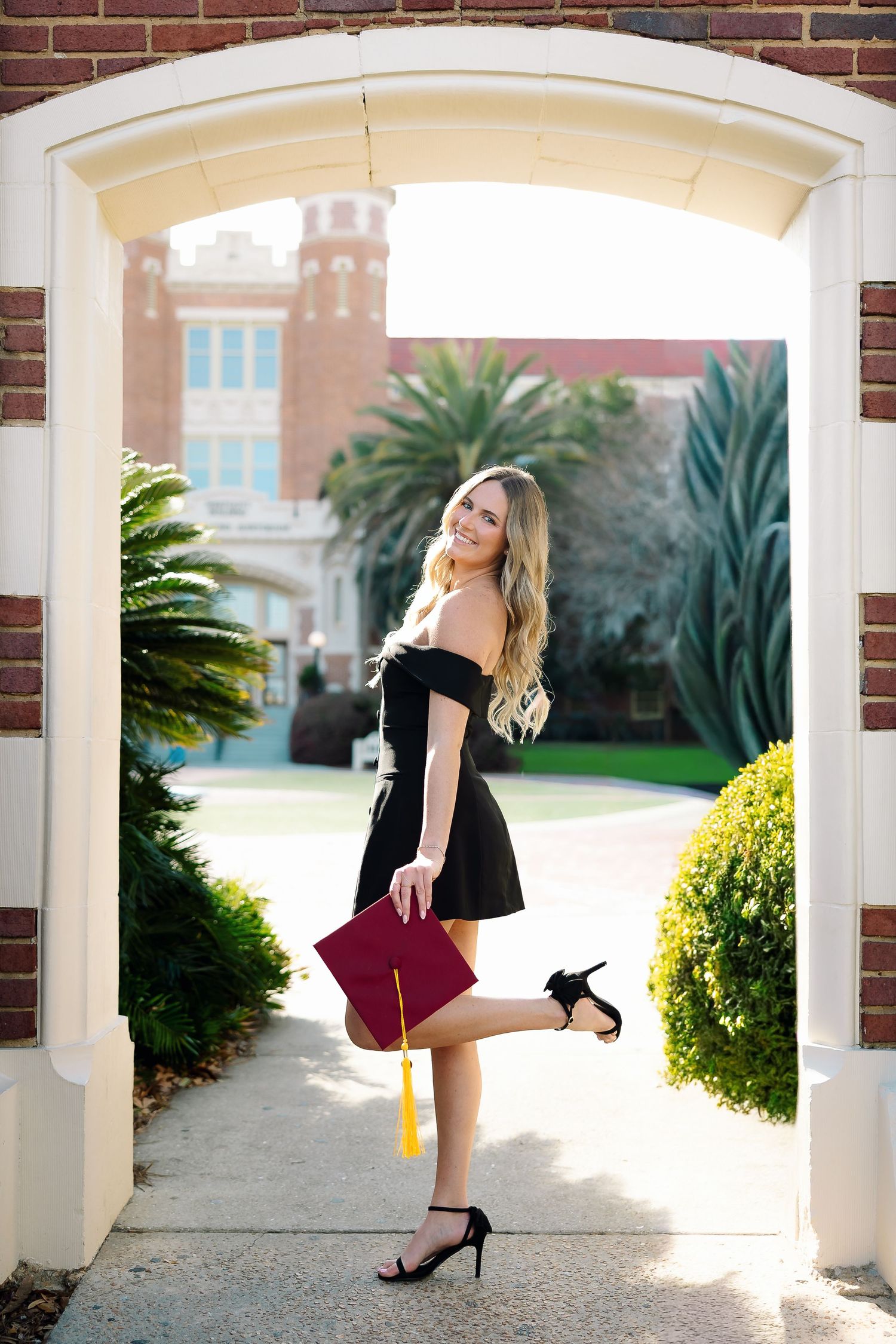 A woman in a black dress poses elegantly under a white columned archway with palm trees and brick walls in the background.