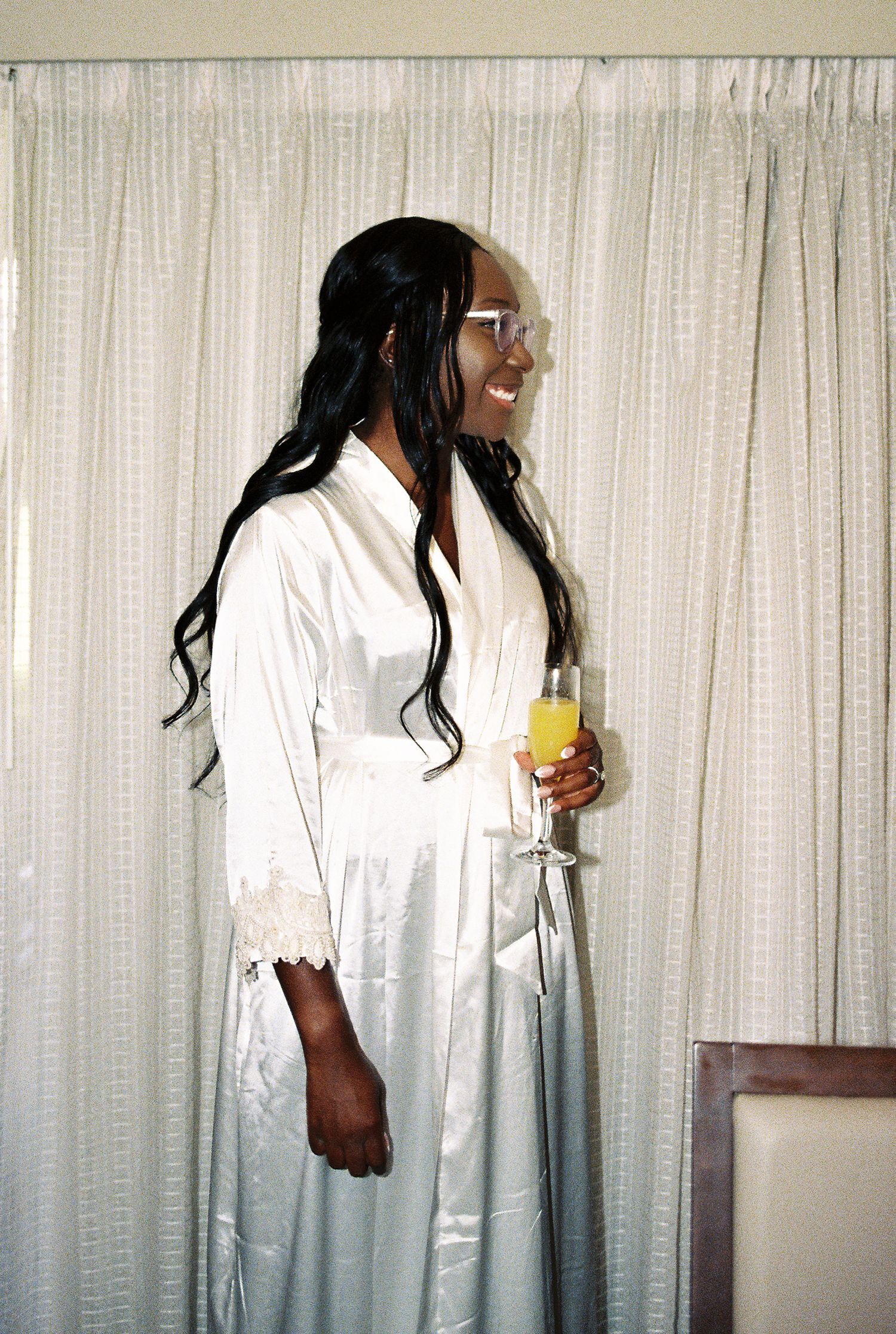 Smiling bride in white satin robe holds champagne glass while standing before sheer curtains.