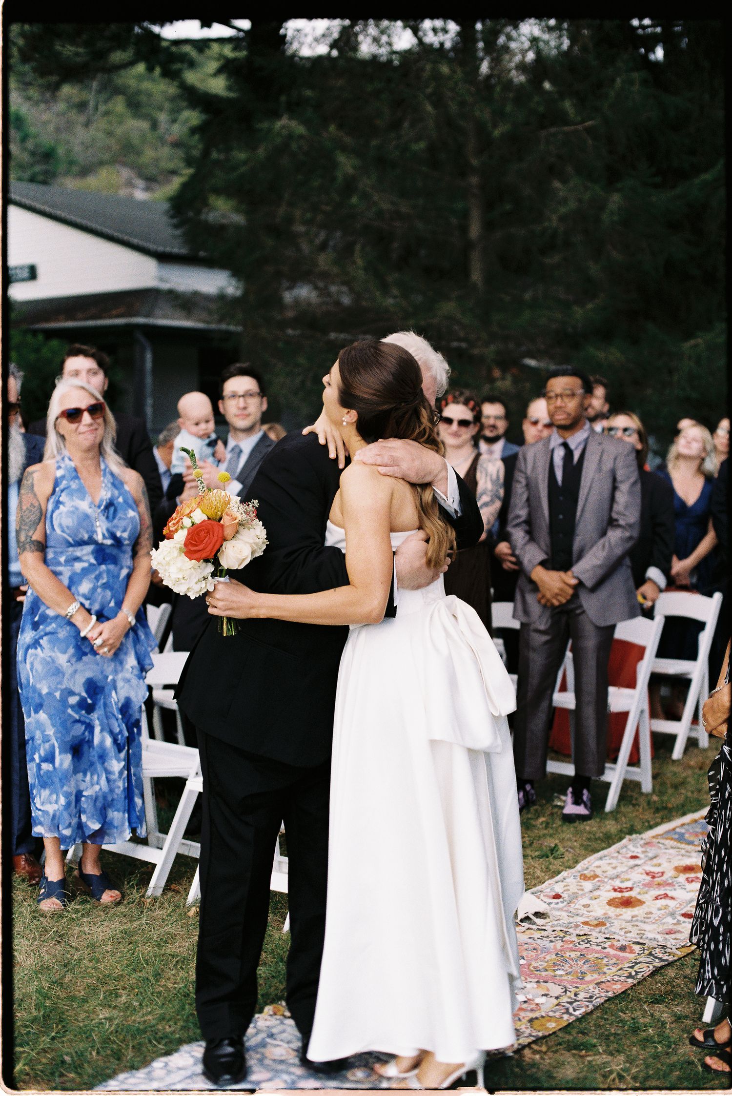 Bride and groom embrace at outdoor wedding ceremony aisle, guests watching as bride holds colorful bouquet on floral runner.