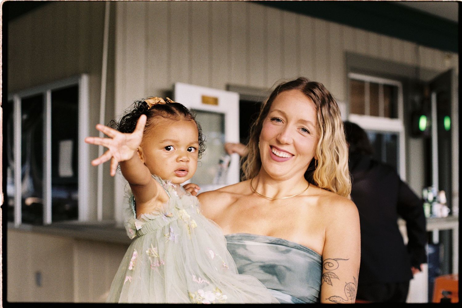A smiling woman in a strapless dress holds a young girl in a sparkly outfit at an outdoor wedding.