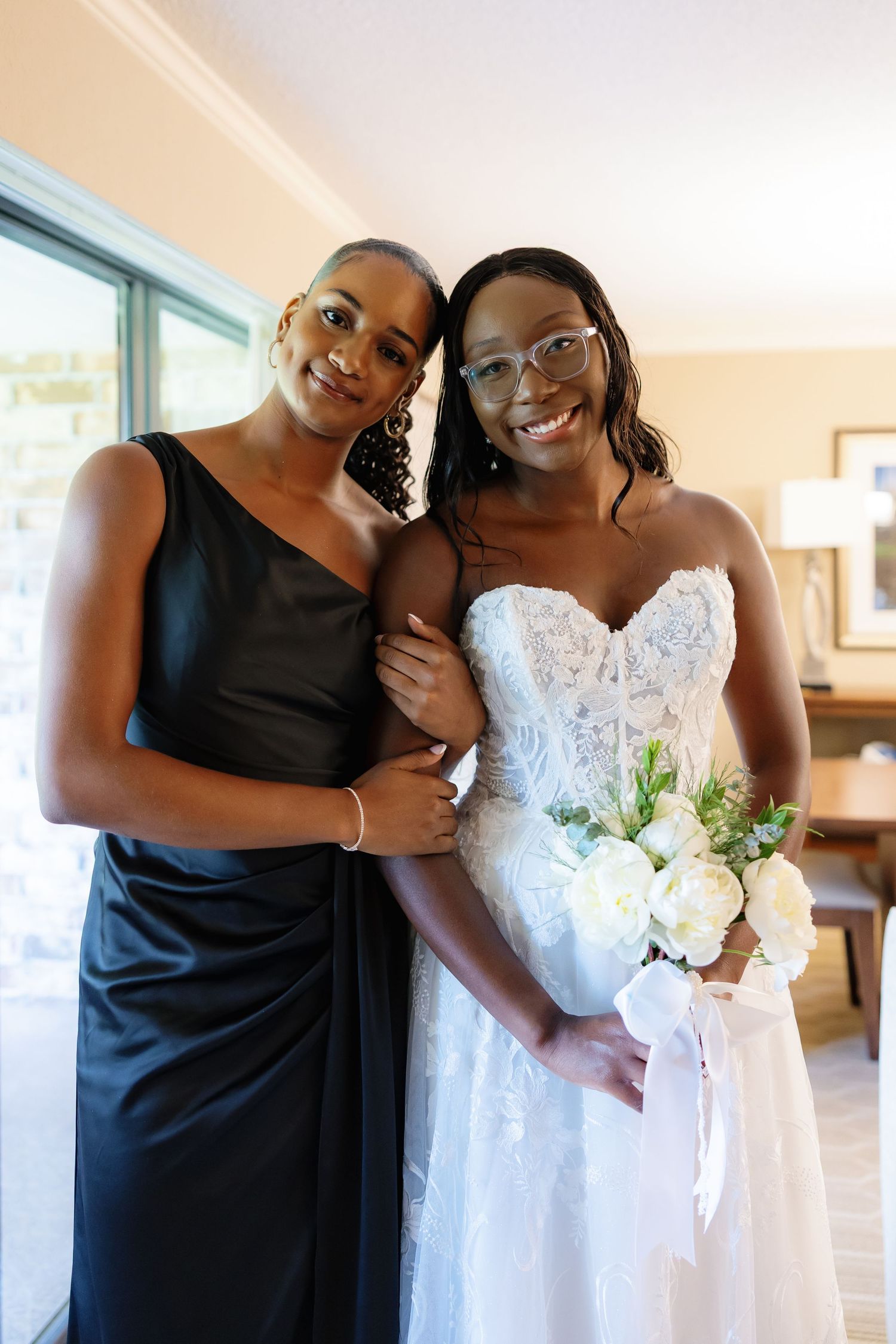 Smiling bride in strapless lace gown holding white peonies poses with bridesmaid in black dress indoors.