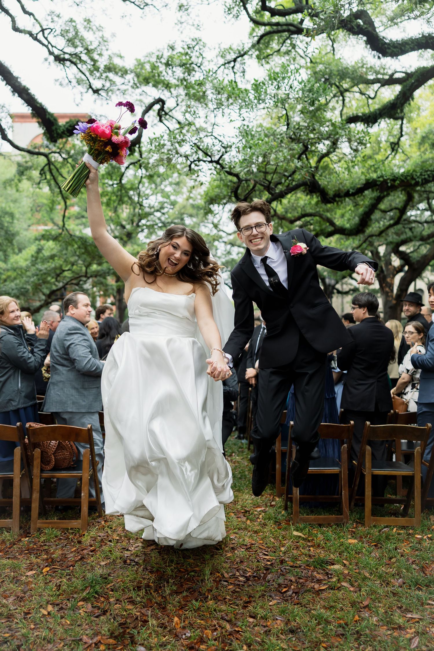 Joyful newlyweds jumping and holding hands while walking down the aisle at an outdoor wedding ceremony.