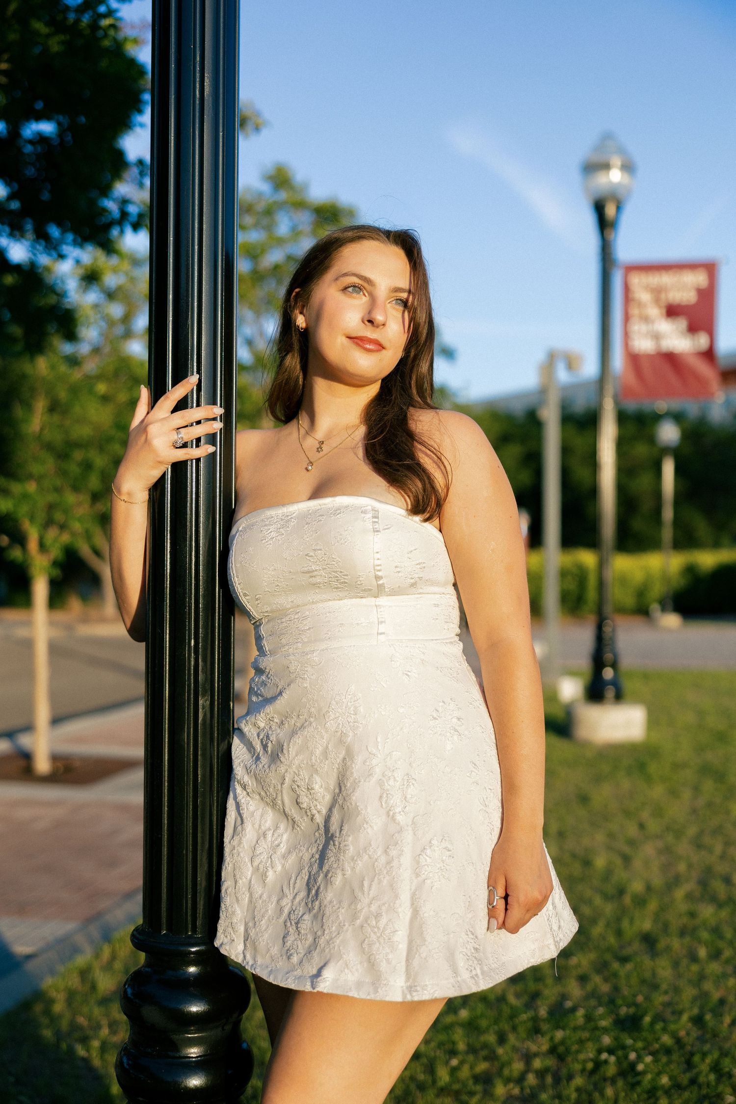 Young woman in white strapless mini dress posing by a black lamp post in a sunny park setting.
