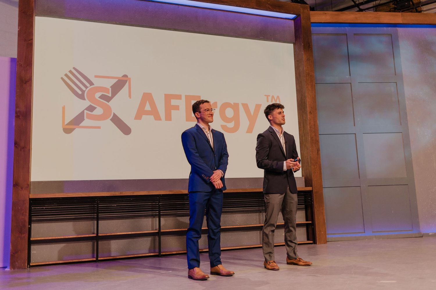 People presenting on stage at a business conference with projection screens and purple accent lighting.