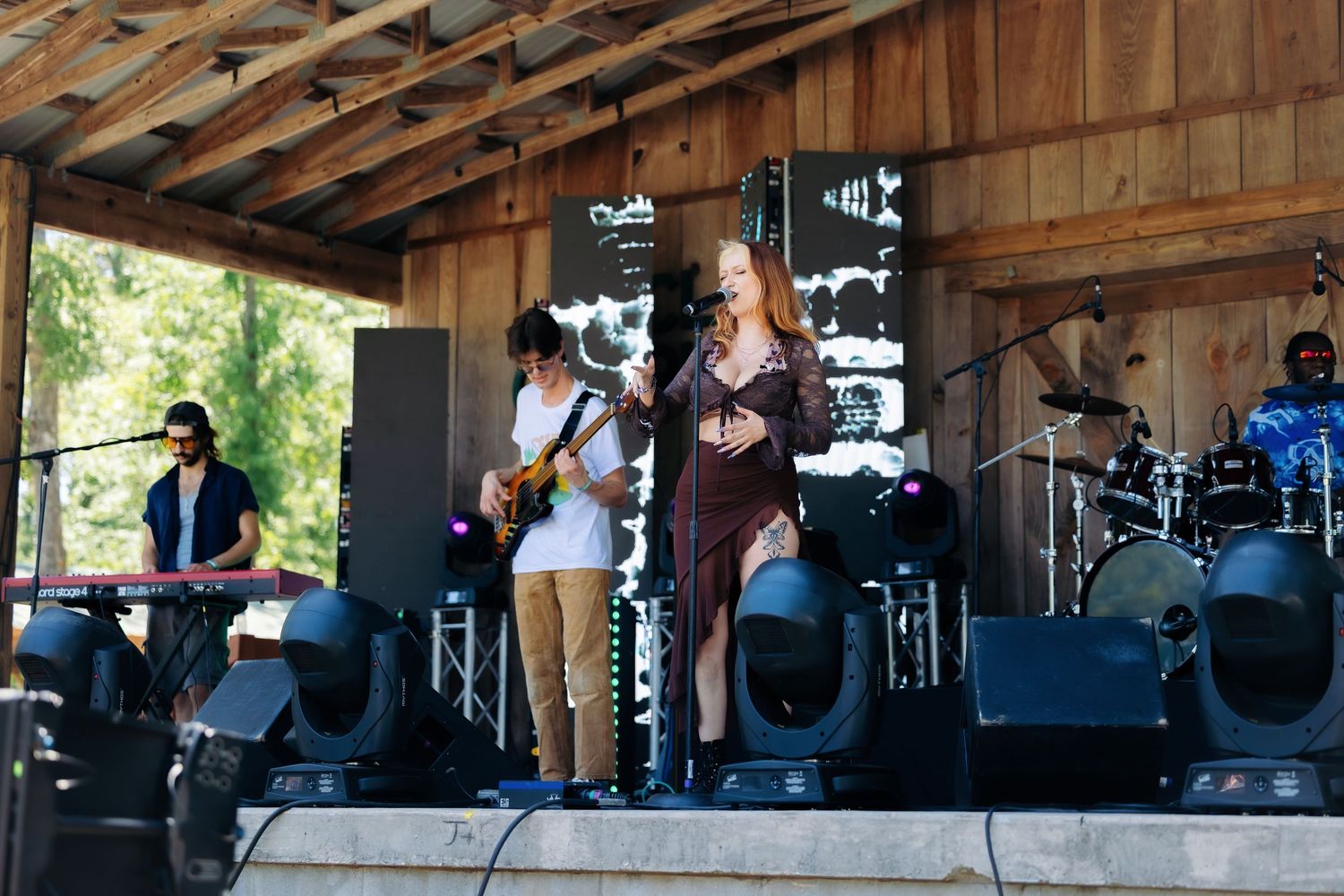 Musicians perform on an outdoor stage with wooden beams overhead during a daytime concert.