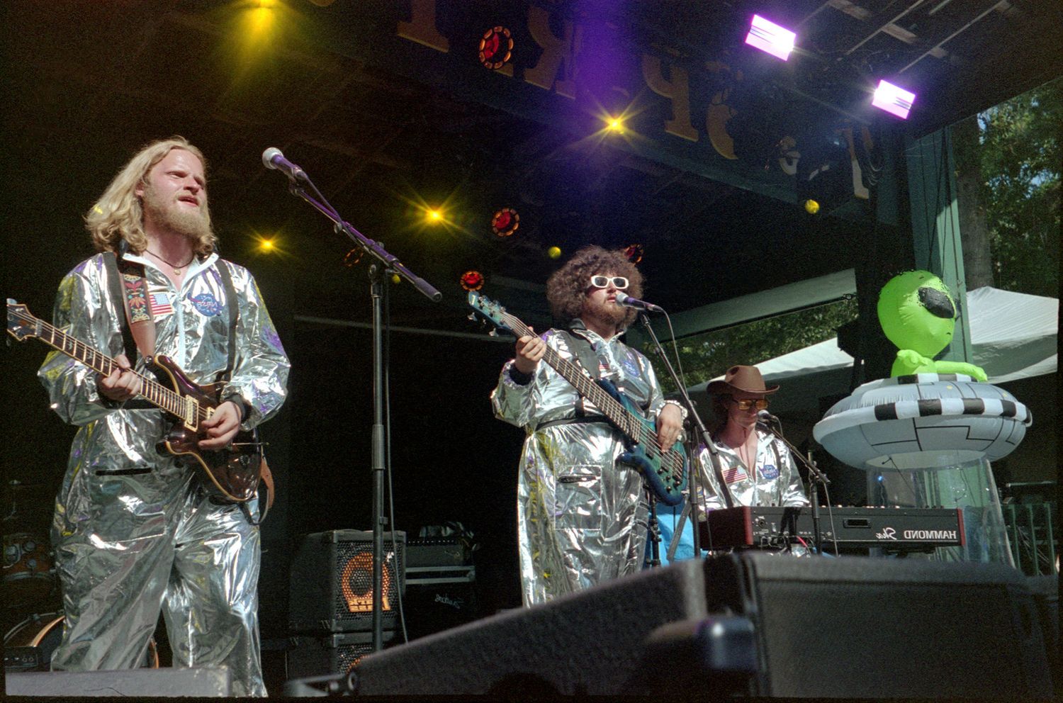 Musicians perform on stage wearing matching silver metallic outfits under colorful stage lighting at a concert performance.