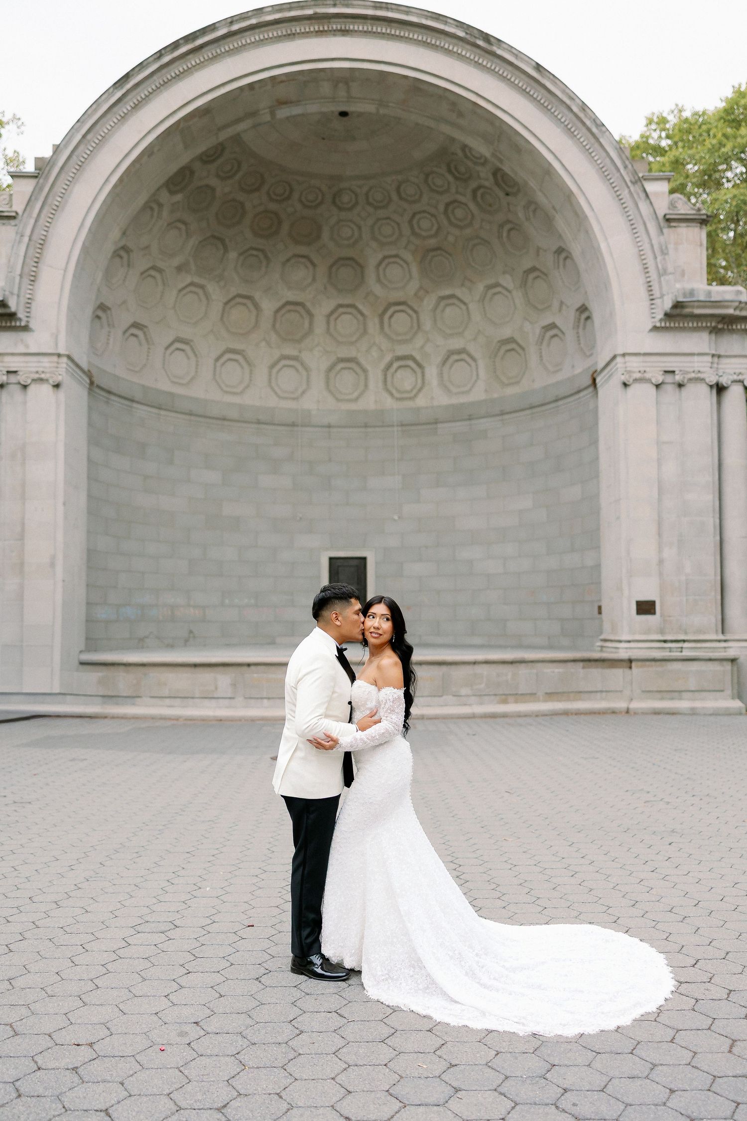A couple in formal wedding attire shares romantic moments in front of an ornate stone archway with domed ceiling.